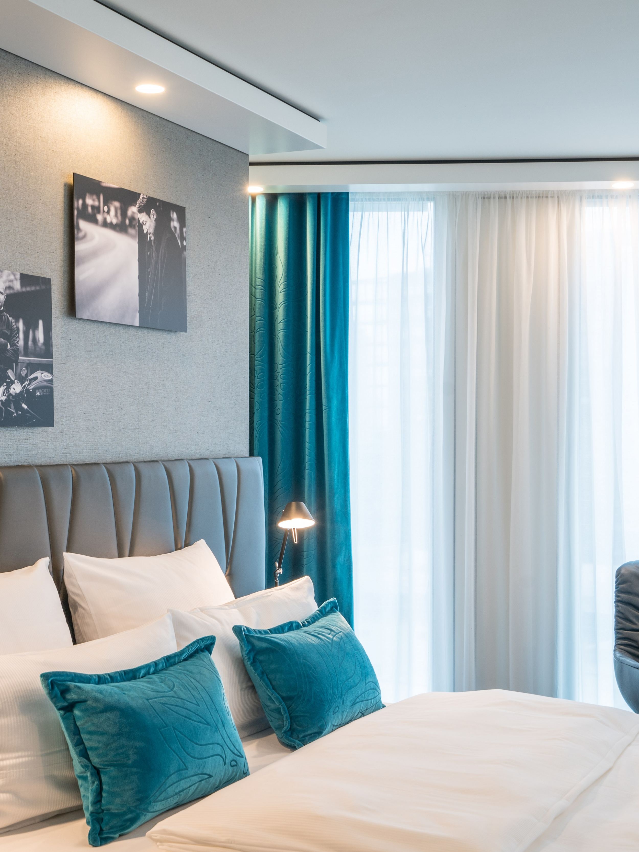 Designhotel Berlin Upper West Motel One seitlicher Blick in ein helles Zimmer mit komfortablem Doppelbett, einem Stuhl mit Tisch und Stehlampe vor großer Fensterfront mit Vorhängen sowie drei Bilder an der Wand