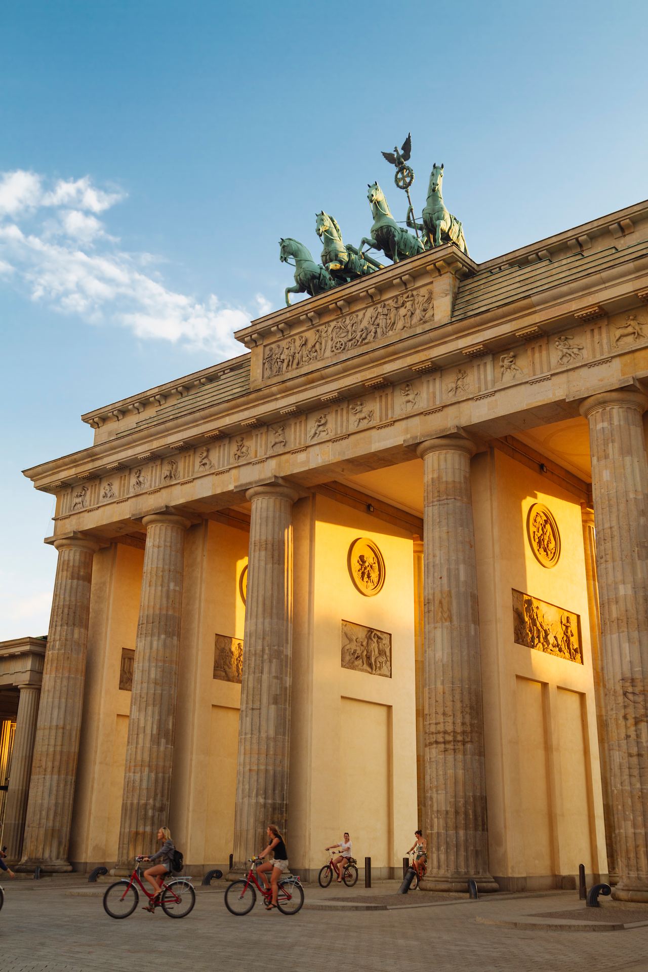 Brandenburger Tor in Berlin bei Sonnenuntergang, beleuchtet, mit mehreren Radfahrern vor den hohen Säulen und der Quadriga auf dem Dach.