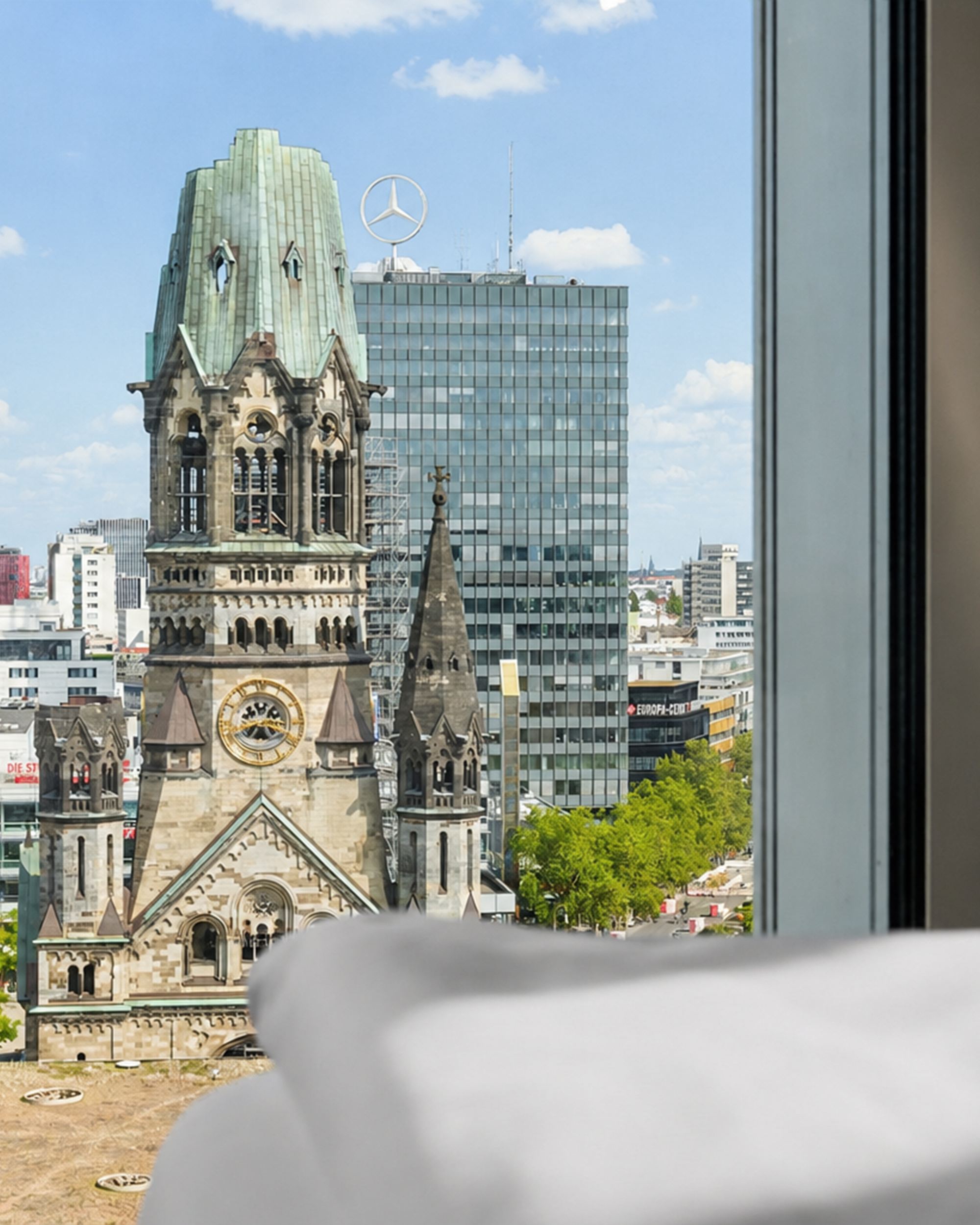 Blick aus einem Hotelzimmerfenster auf die Kaiser-Wilhelm-Gedächtniskirche und moderne Gebäude in der Berliner Innenstadt bei klarem Himmel.