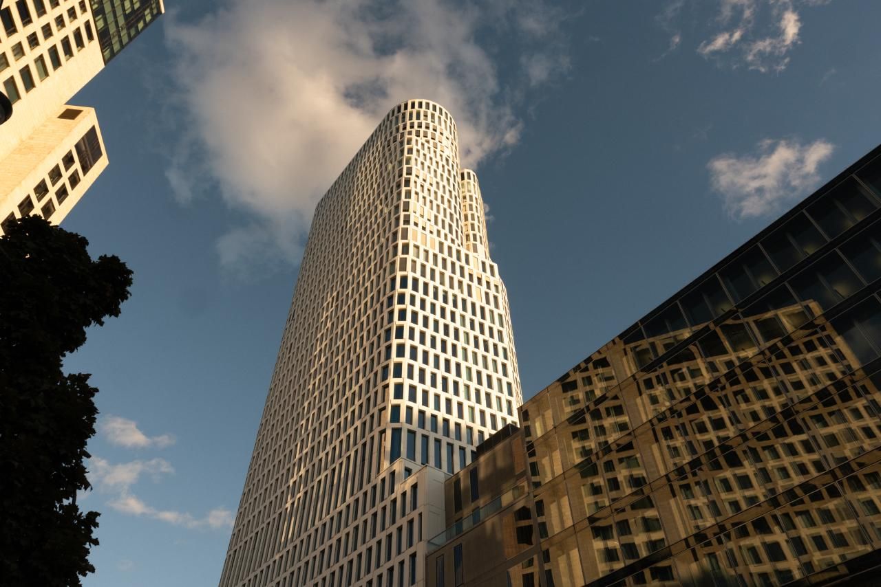 Moderner Wolkenkratzer mit wellenförmiger Fassade, aufgenommen bei warmem Tageslicht, vor blauem Himmel mit einigen Wolken, umgeben von weiteren Gebäuden. Ideal für Geschäftsreisende, die urbanes Ambiente und zeitgemäße Architektur schätzen.