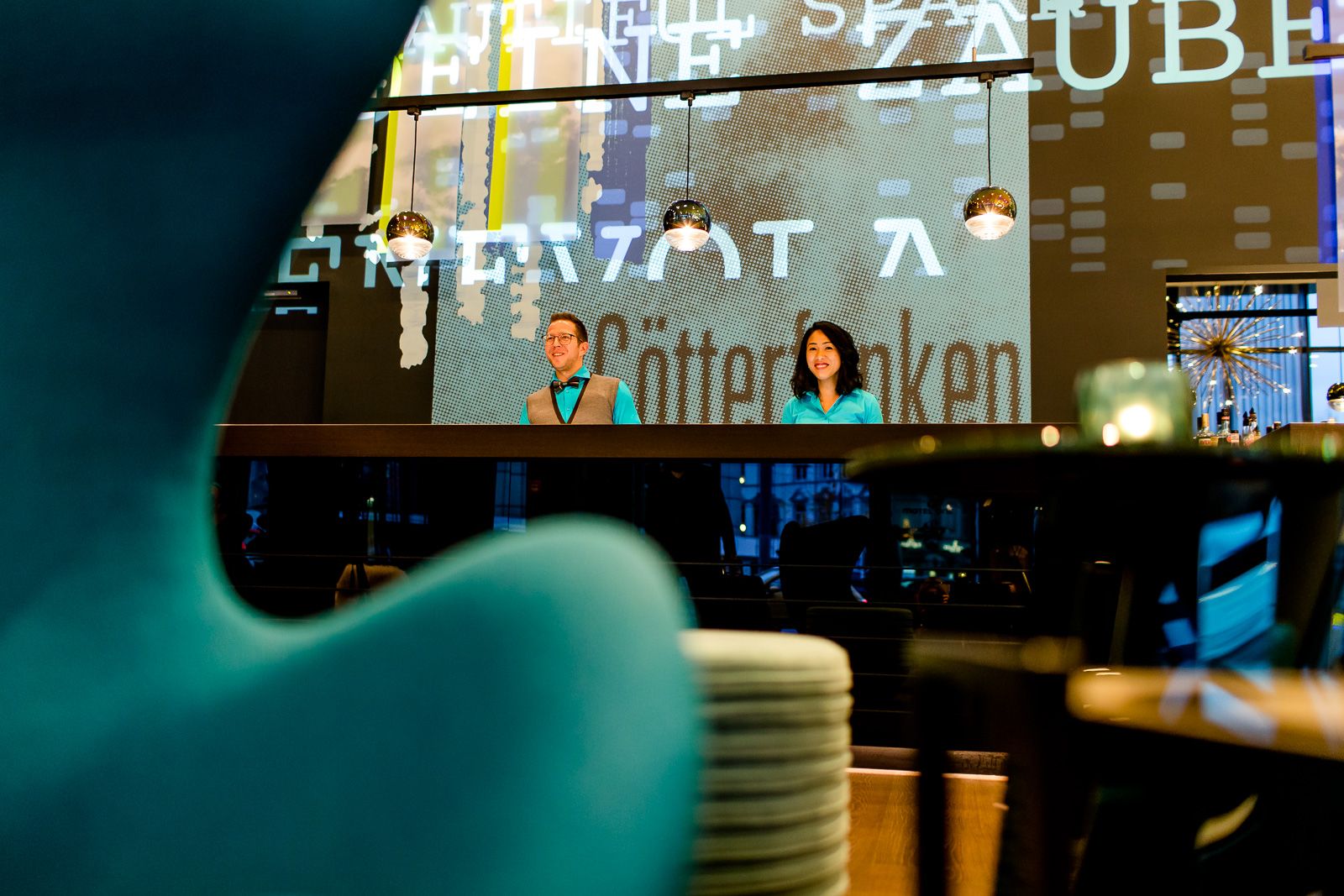 Designhotel Bonn Beethoven Motel One Rezeption mit zwei Mitarbeitenden sowie Anschnitt eines Egg Chairs im Vordergrund