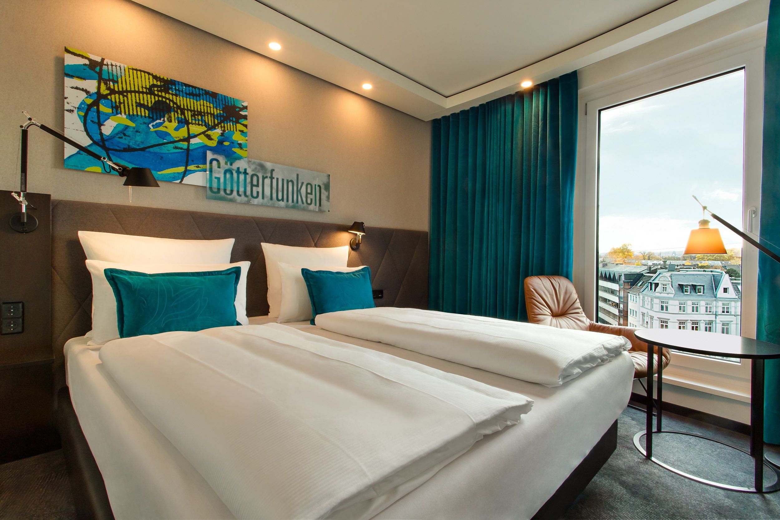 hotel Bonn Beethoven Motel One bright room with a large double bed and a table and chair in front of a large window overlooking the rooftops of the city