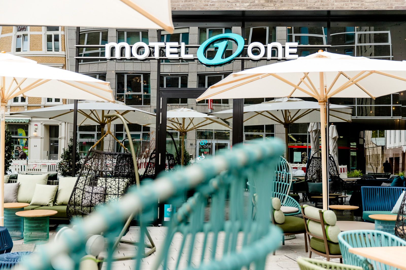 Hotel Bonn Hauptbahnhof Motel One Terrasse mit mehreren Sitzgelegenheiten sowie Blick auf die verspiegelte Fensterfront mit Logo