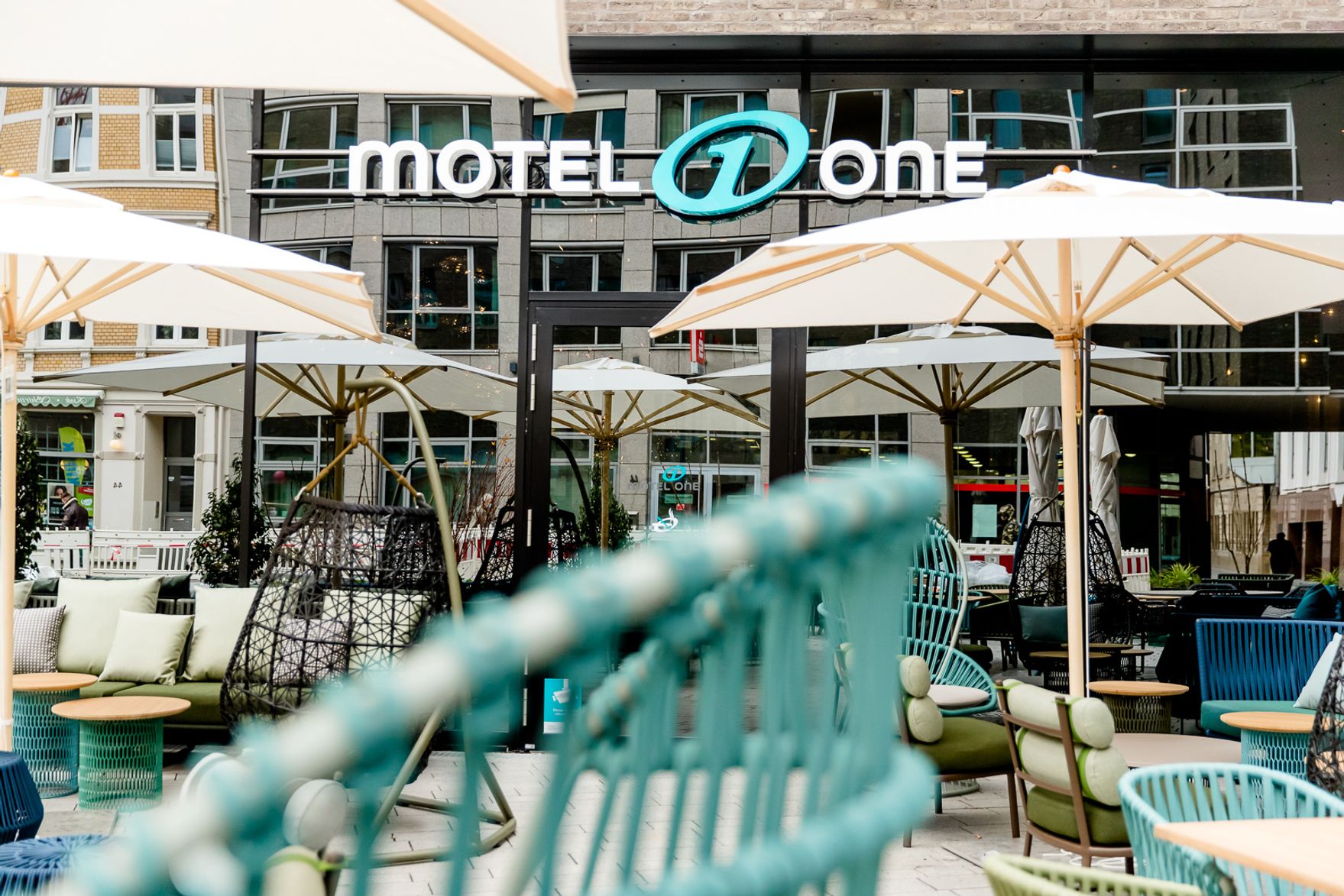 Hotel Bonn Hauptbahnhof Motel One Terrasse mit mehreren Sitzgelegenheiten sowie Blick auf die verspiegelte Fensterfront mit Logo