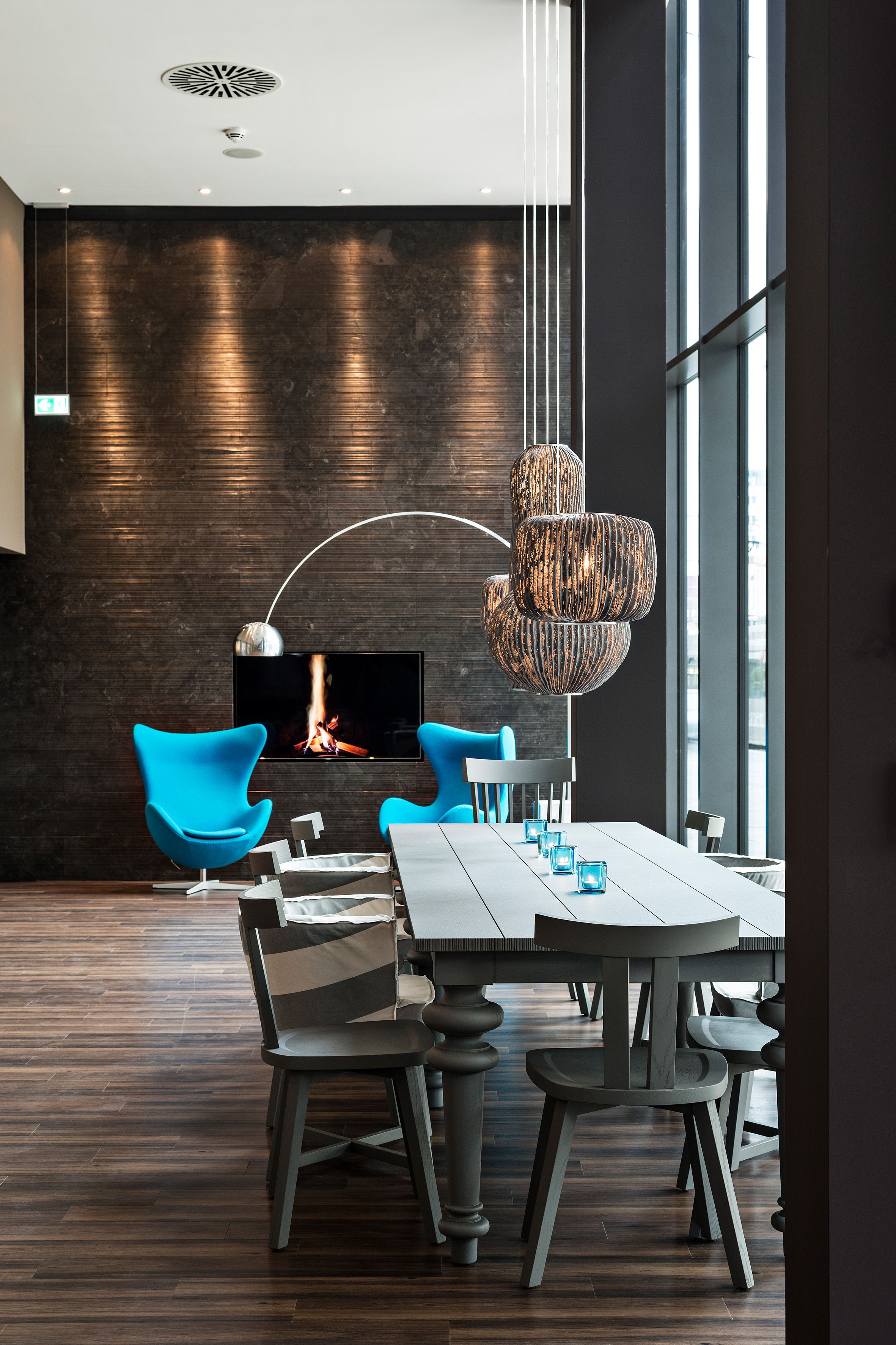 Designhotel Bremen Motel One helle Lounge mit langem Tisch sowie zwei Egg Chairs im Hintergrund mit Fernseher an der Wand