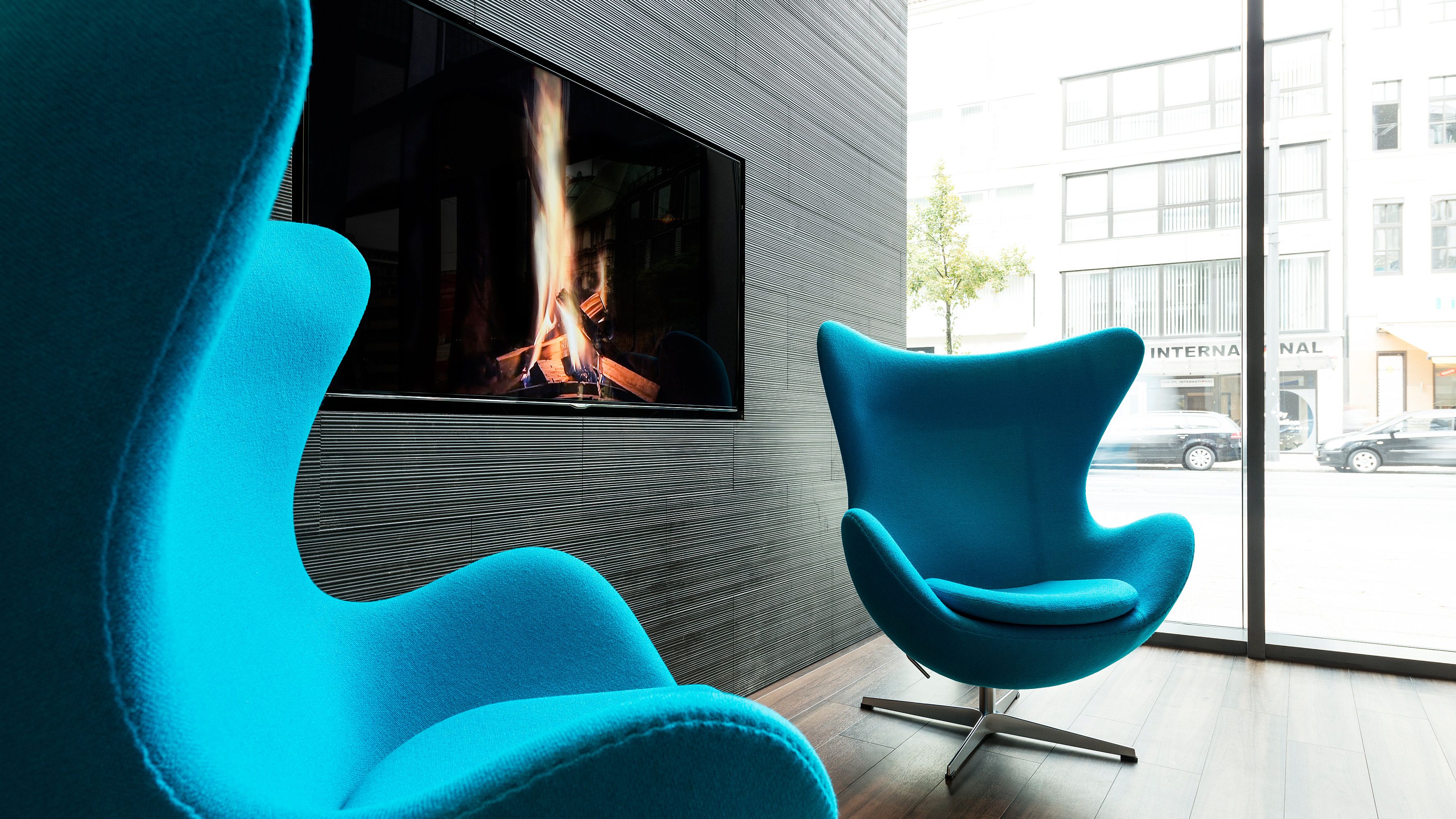 Hotel Bremen Motel One zwei Egg Chairs vor bodentiefem Fenster mit Fernseher an der Wand, der ein brennendes Feuer zeigt