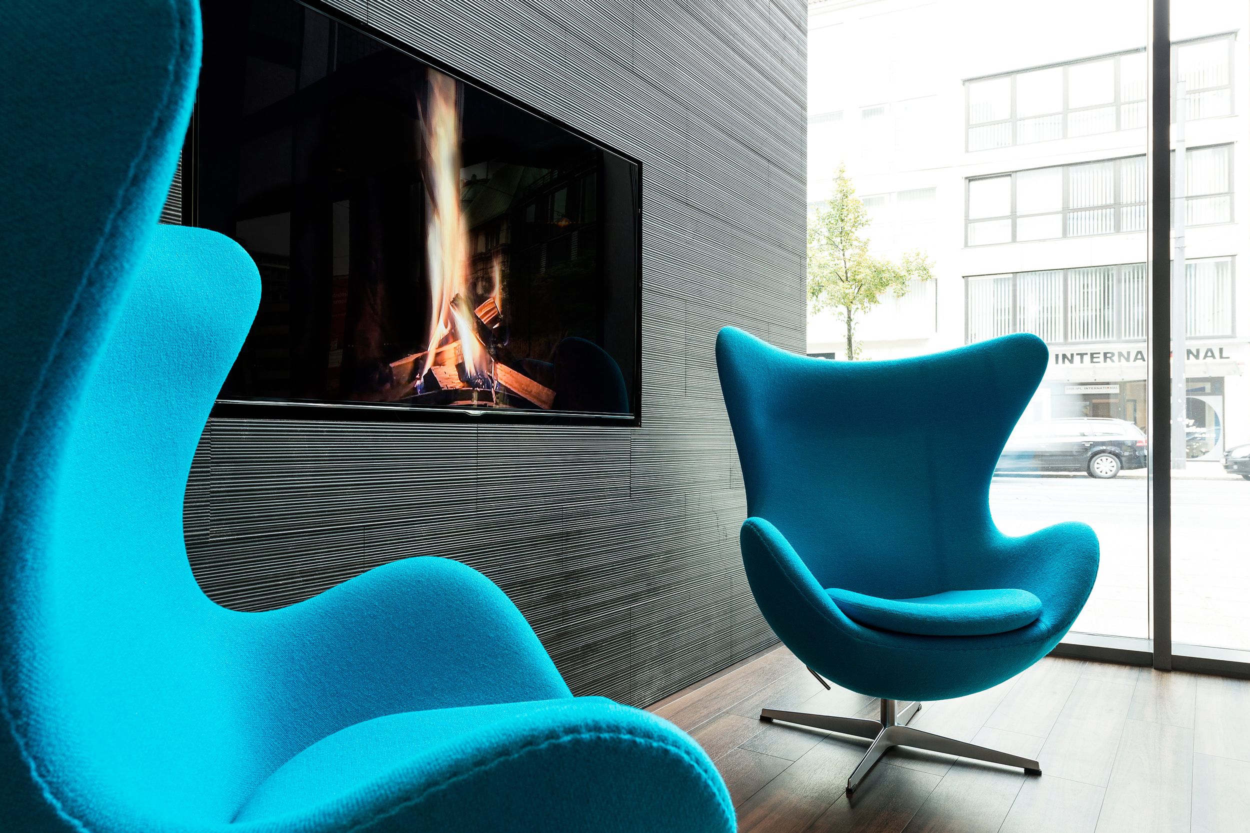 hotel Bremen Motel One two egg chairs in front of a floor-to-ceiling window with a television on the wall showing a burning fire
