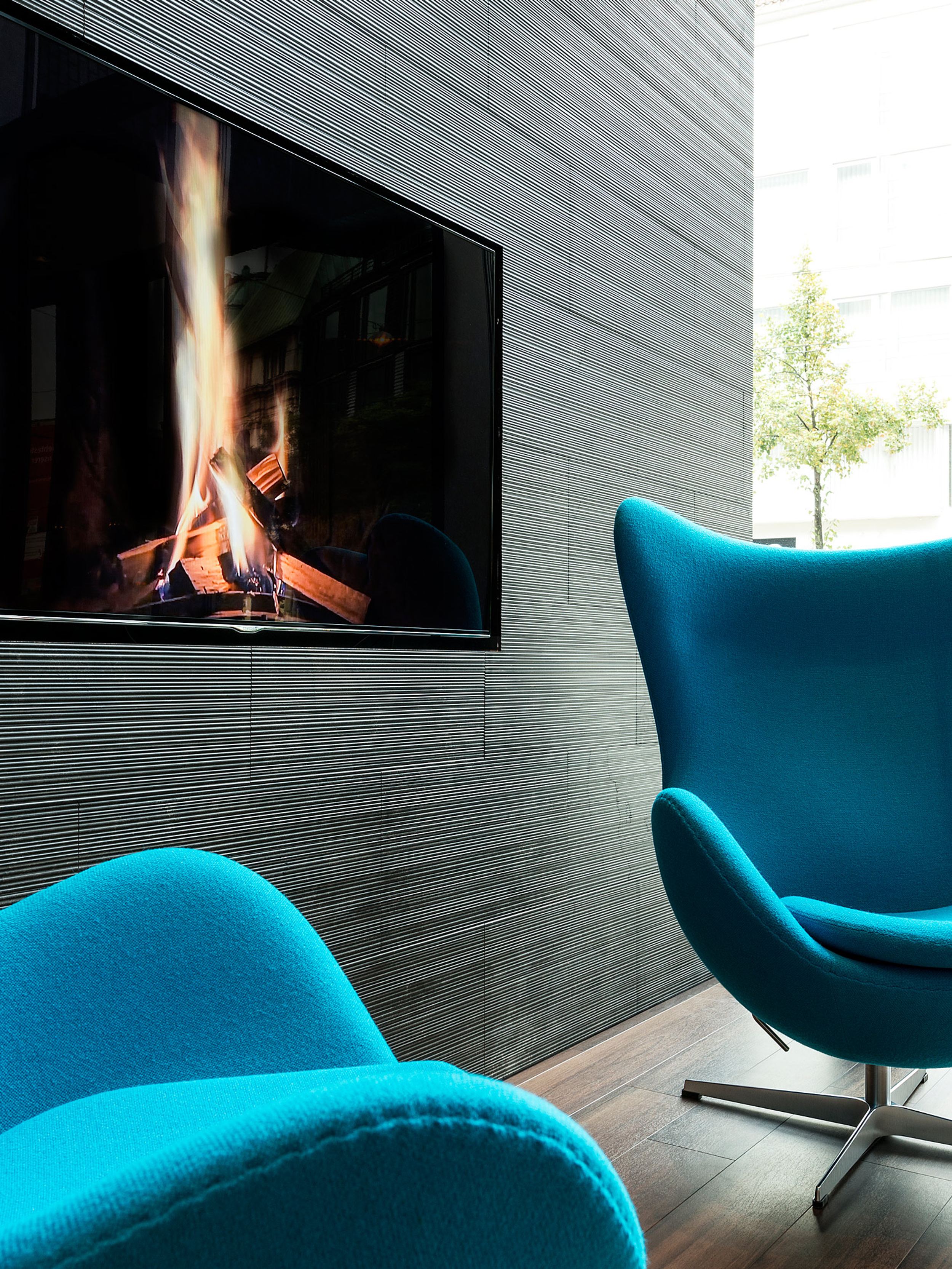 hotel Bremen Motel One two egg chairs in front of a floor-to-ceiling window with a television on the wall showing a burning fire