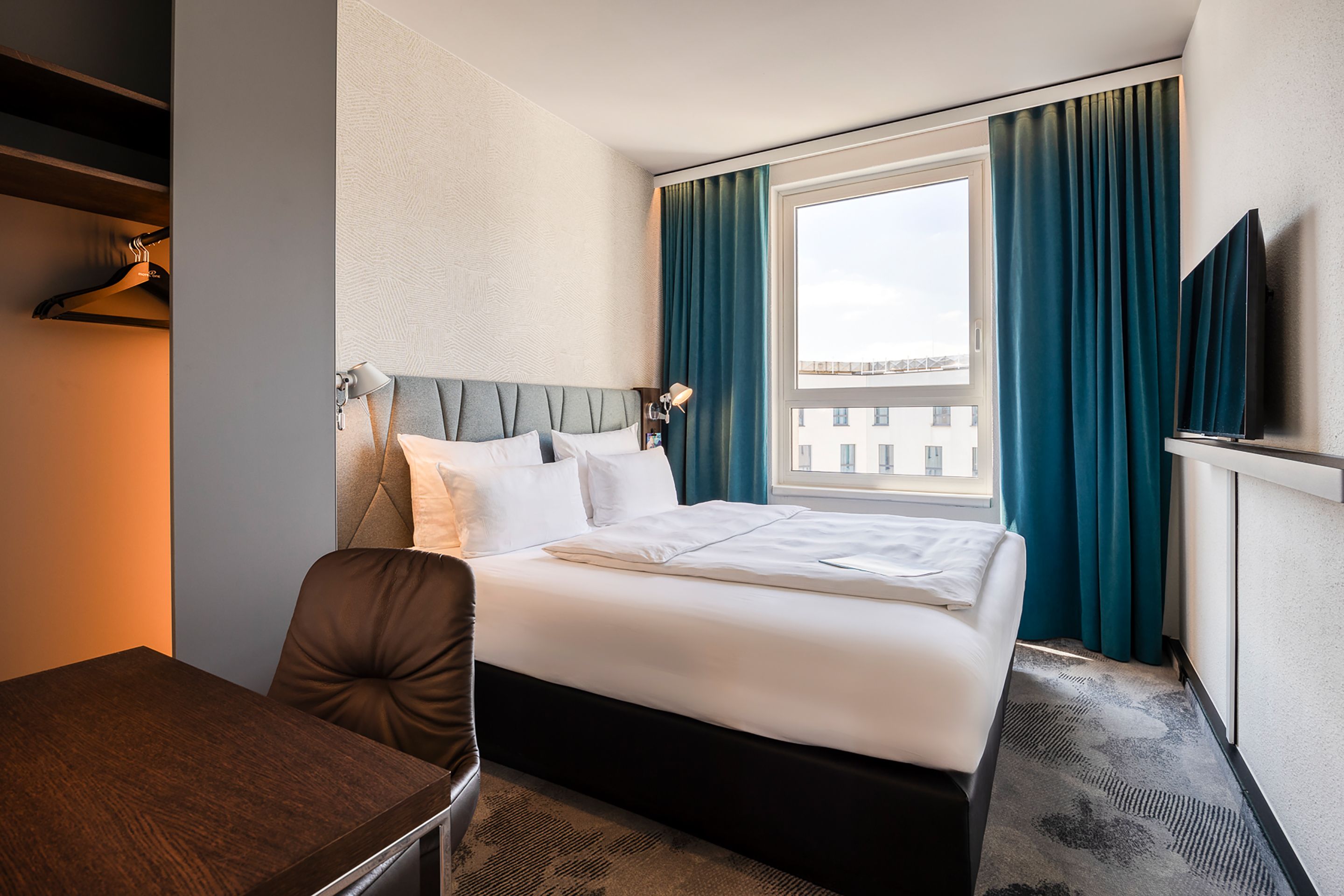 Designhotel Brüssel Motel One seitlicher Blick auf großes Doppelbett sowie ein Fenster mit zwei Vorhängen in türkis