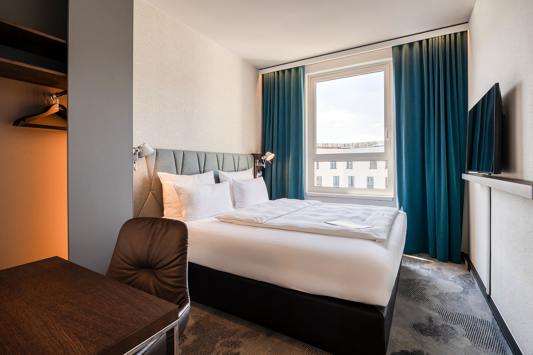 Designhotel Brüssel Motel One seitlicher Blick auf großes Doppelbett sowie ein Fenster mit zwei Vorhängen in türkis