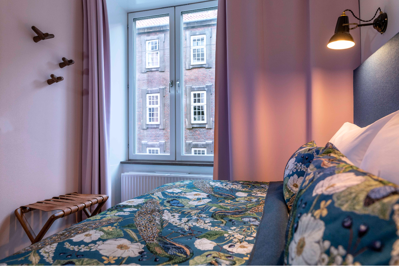 Designhotel Kopenhagen Motel One Zimmer mit gemütlichem Bett und gemusterter Bettwäsche neben großem Fenster mit Vorhängen in rosa