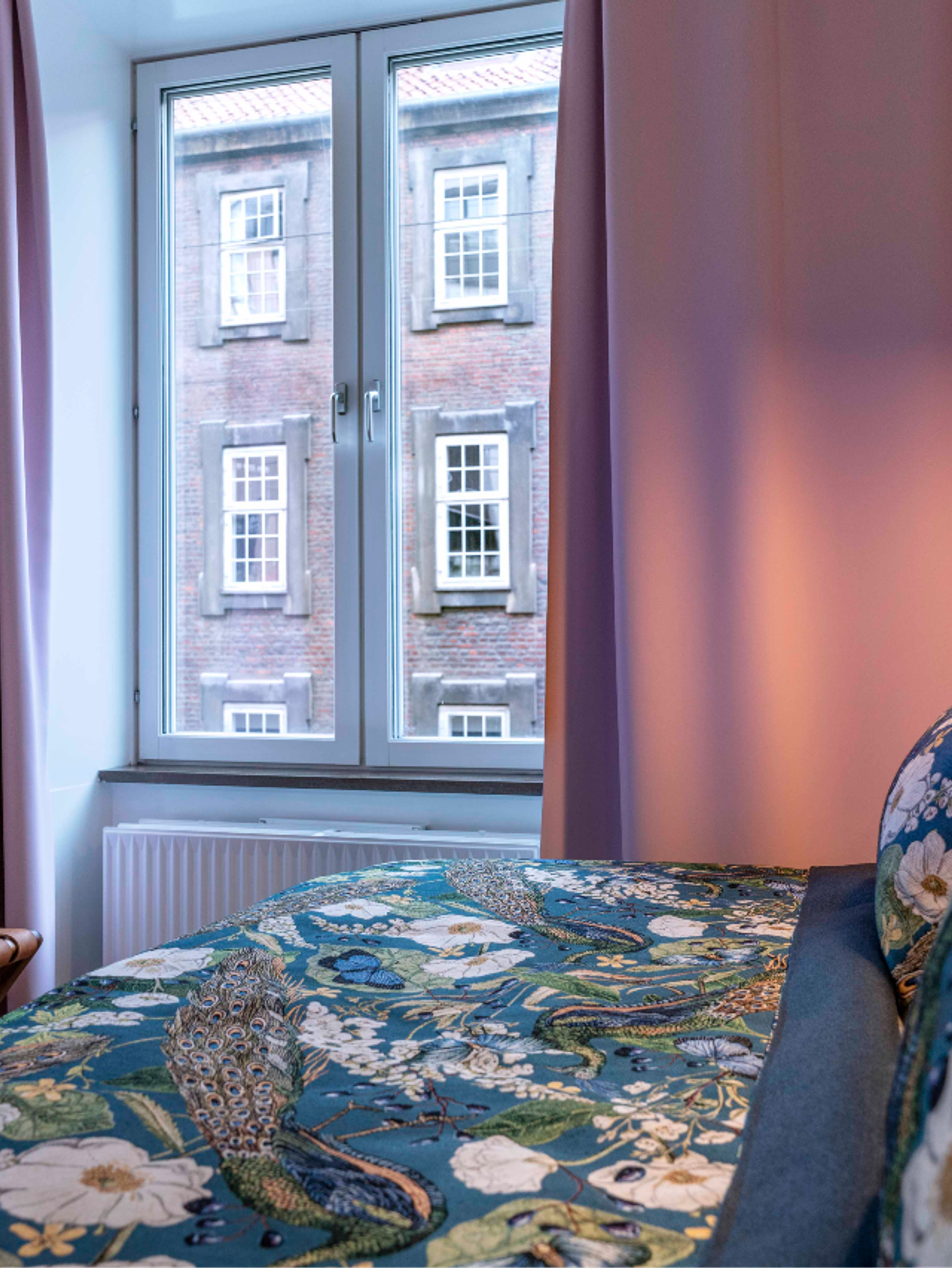 Designhotel Kopenhagen Motel One Zimmer mit gemütlichem Bett und gemusterter Bettwäsche neben großem Fenster mit Vorhängen in rosa