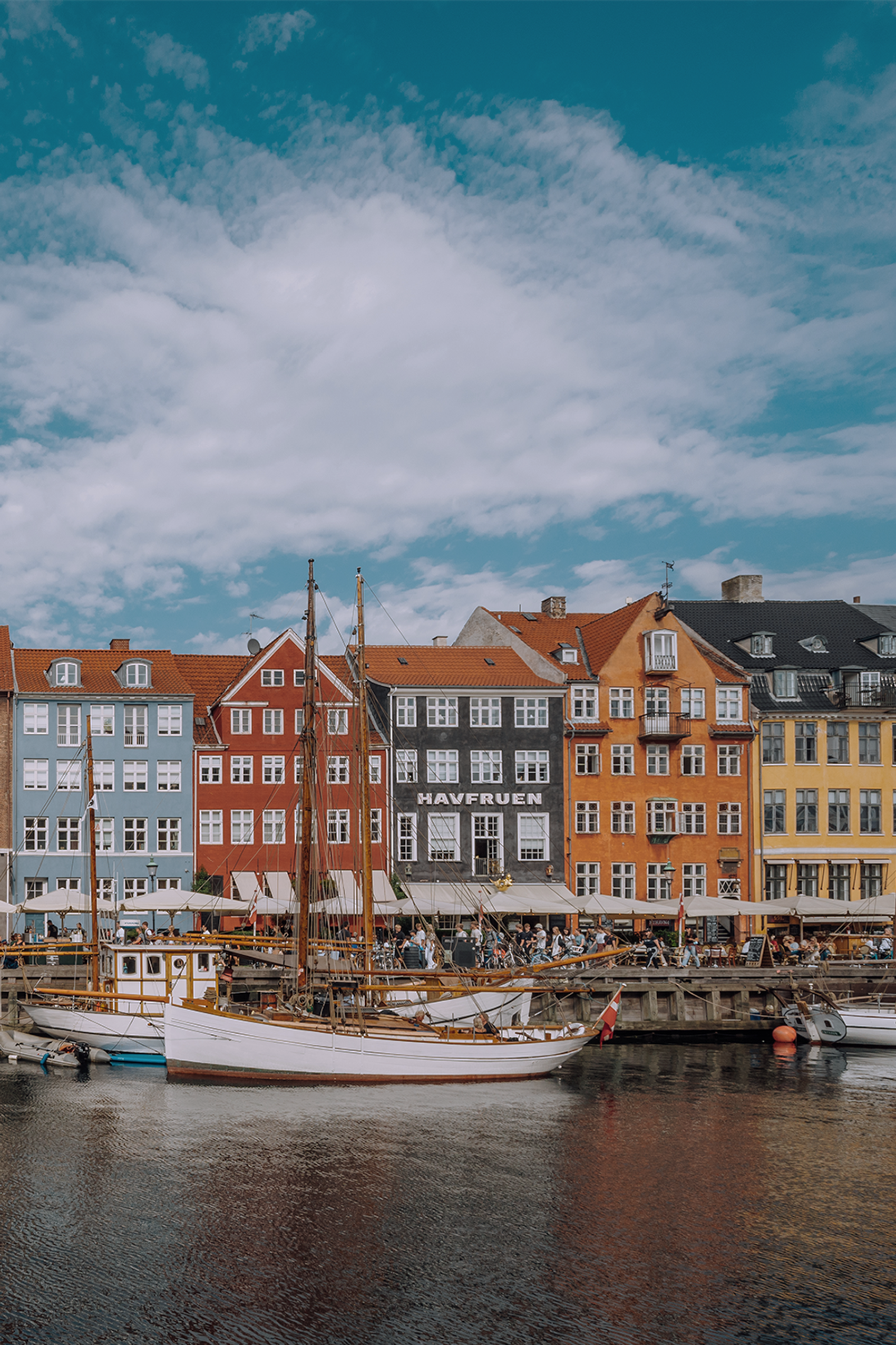 Bunte historische Häuserzeile am Wasser mit kleinen Segelbooten im Vordergrund und einem bewölkten Himmel, typisch für einen lebendigen Hafenkai in einer europäischen Stadt.