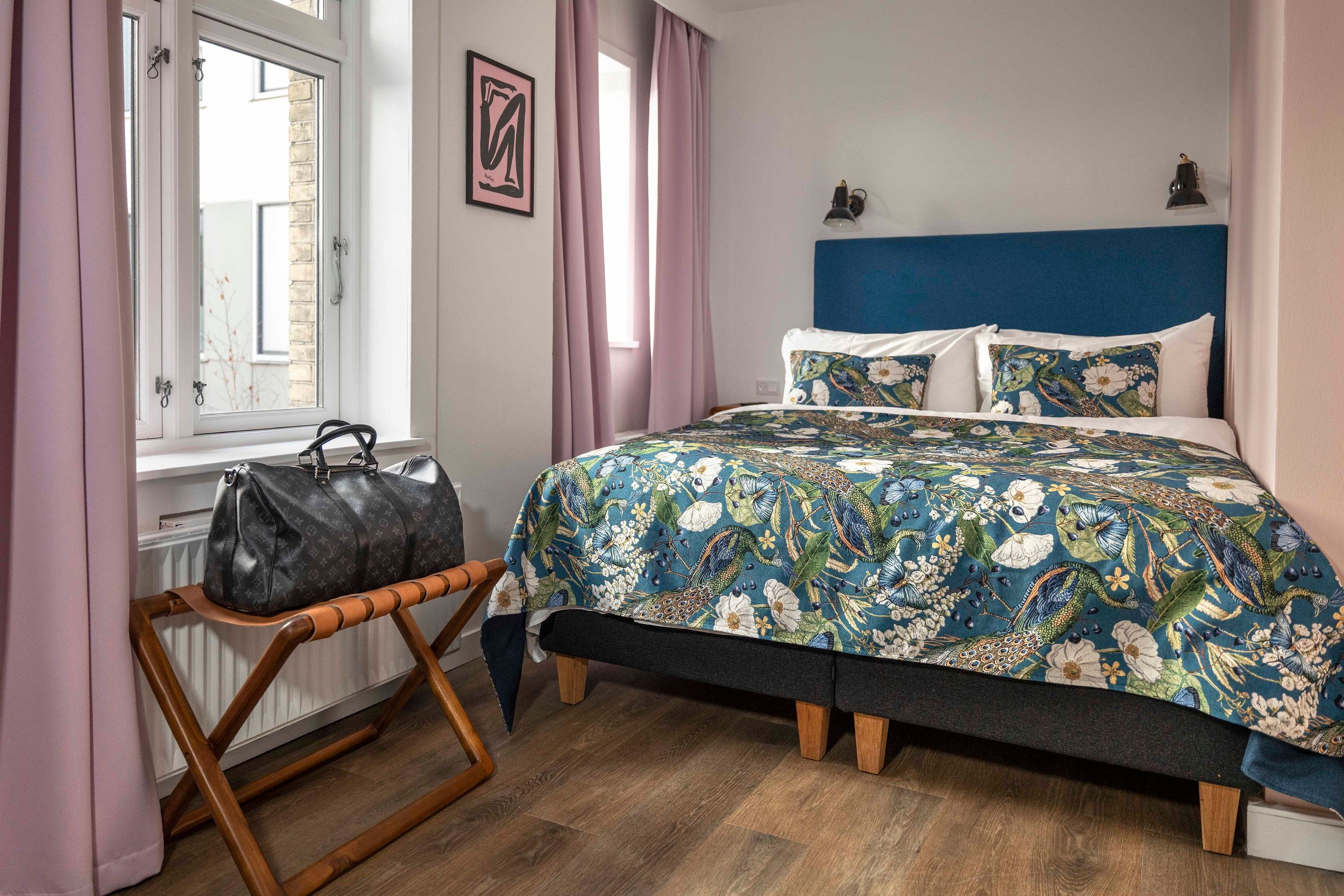 Designhotel Kopenhagen Motel One Blick auf ein gemütliches Doppelbett mit gemusterter Bettwäsche, zwei Fenster mit Vorhängen in rosa sowie eine Tasche auf einer Ablage