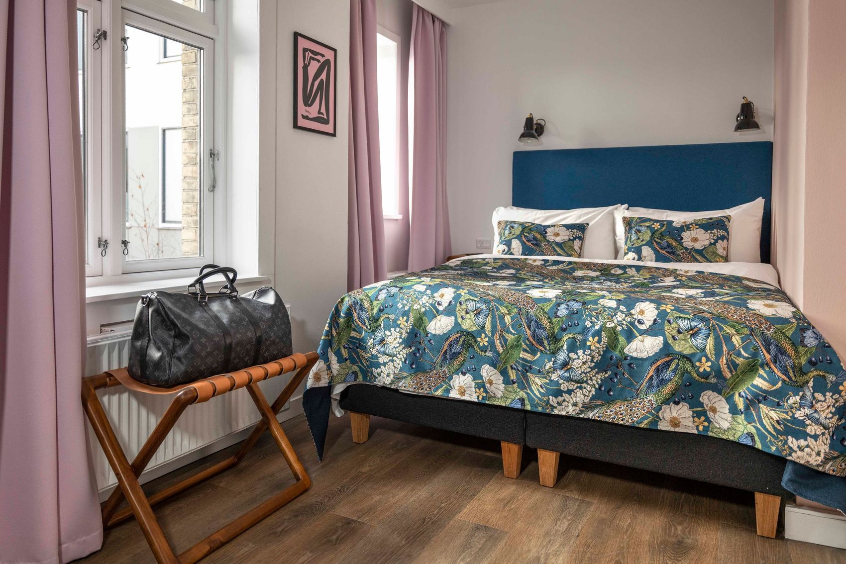 designhotel Copenhagen Motel One view of a cozy double bed with patterned bedding, two windows with pink curtains, and a bag on a shelf