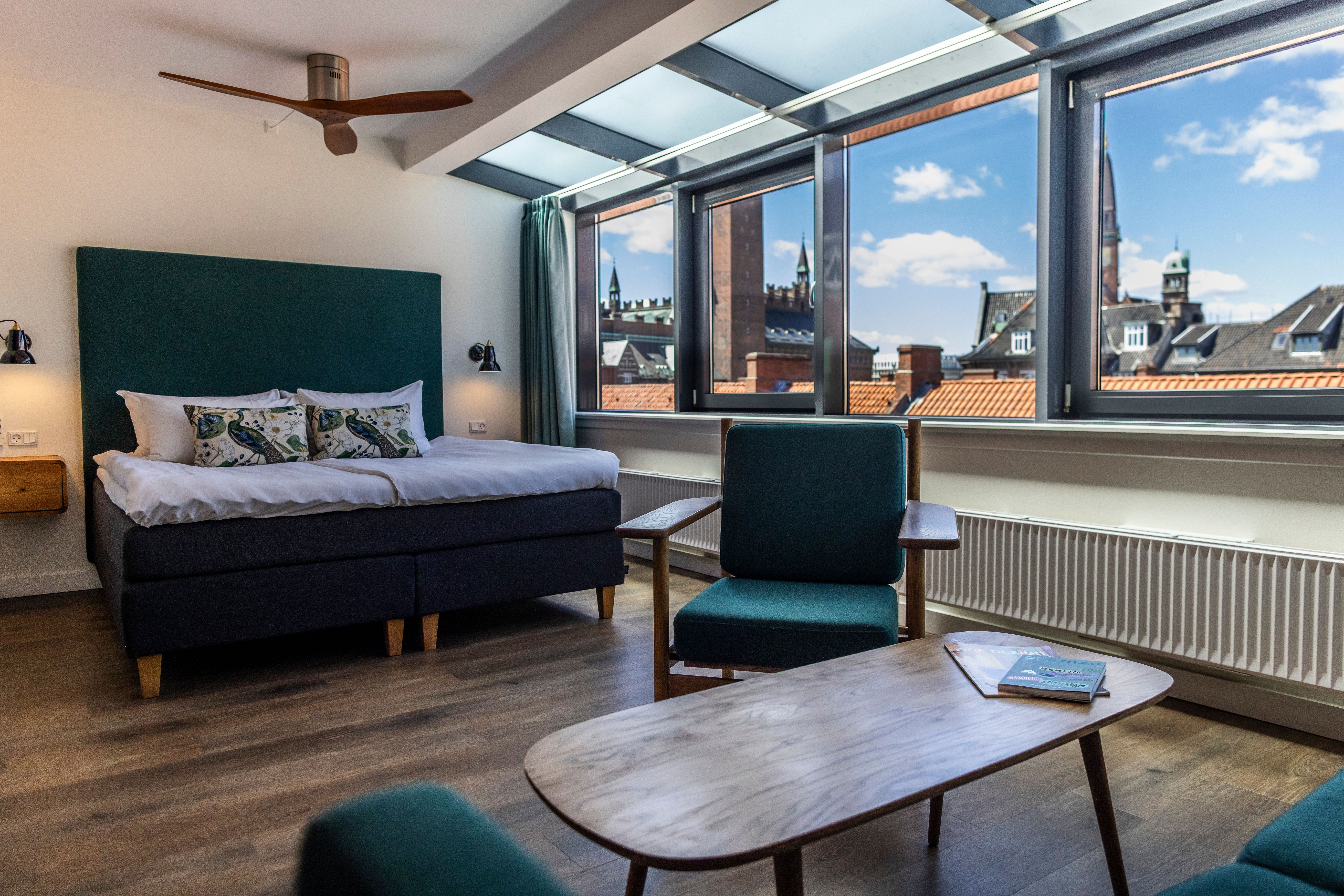Hotel Motel One Copenhagen Modern eingerichtetes Hotelzimmer mit großem Fenster und Blick auf historische Stadtgebäude, komfortables Doppelbett, Sitzbereich mit Stühlen und Holztischen, helle, freundliche Atmosphäre durch Tageslicht.