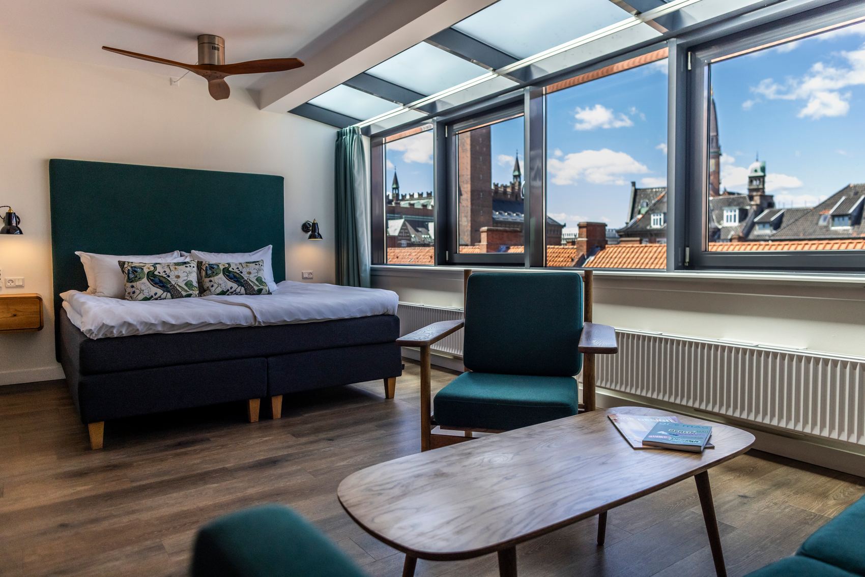 Hotel Motel One Copenhagen Modern eingerichtetes Hotelzimmer mit großem Fenster und Blick auf historische Stadtgebäude, komfortables Doppelbett, Sitzbereich mit Stühlen und Holztischen, helle, freundliche Atmosphäre durch Tageslicht.