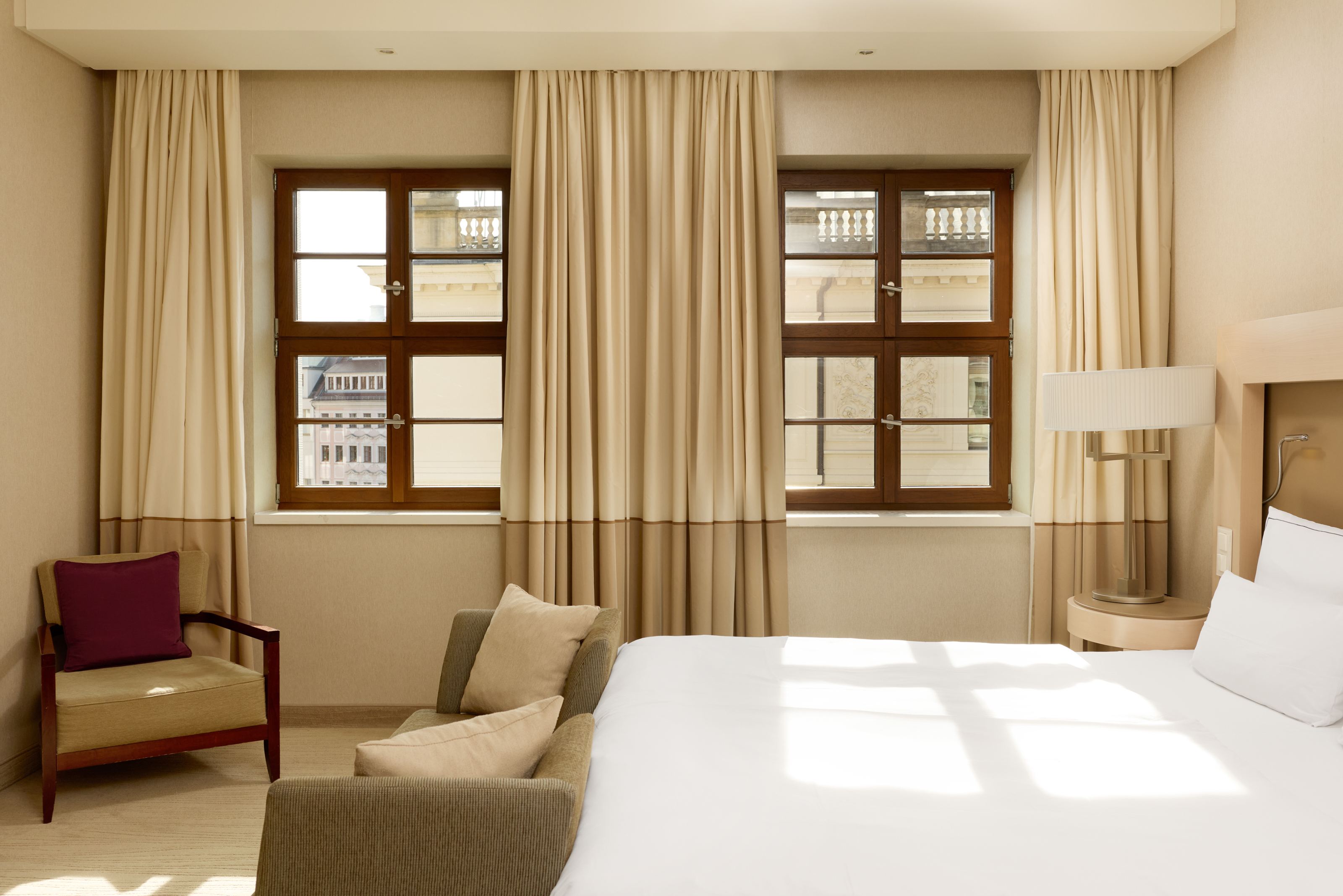 Helles Hotelzimmer mit großen Fenstern, beigen Vorhängen, gemütlichem Bett, einem Sessel und einem Sofa, das zum Entspannen einlädt. Natürliches Licht durchflutet den Raum und schafft eine freundliche, ruhige Atmosphäre. Helles Hotelzimmer mit großen Fenstern, beigen Vorhängen, gemütlichem Bett, einem Sessel und einem Sofa, das zum Entspannen einlädt. Natürliches Licht durchflutet den Raum und schafft eine freundliche, ruhige Atmosphäre.