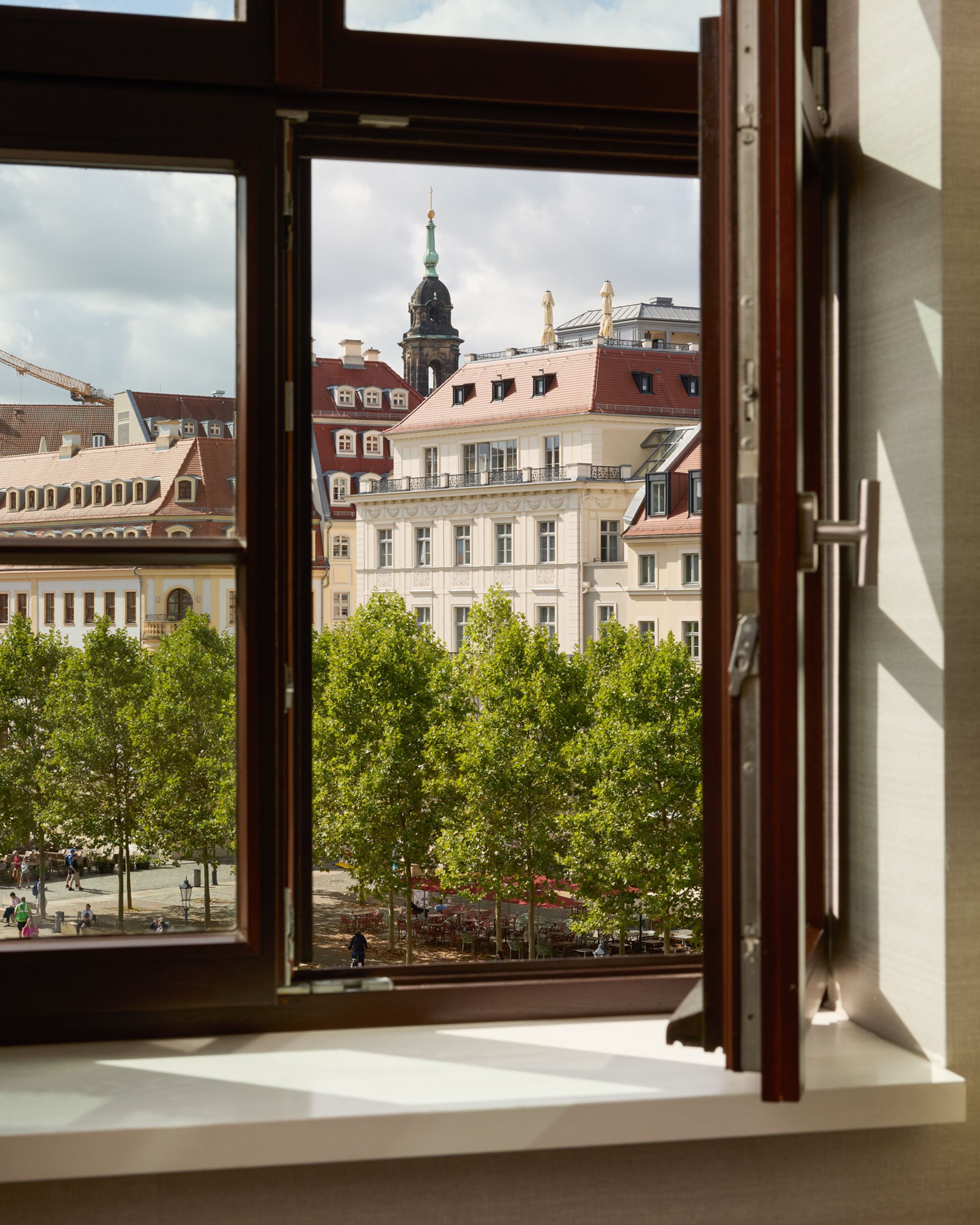 Blick aus offenem Hotelzimmerfenster auf historische Gebäude und grüne Bäume an einem städtischen Platz bei Tageslicht.