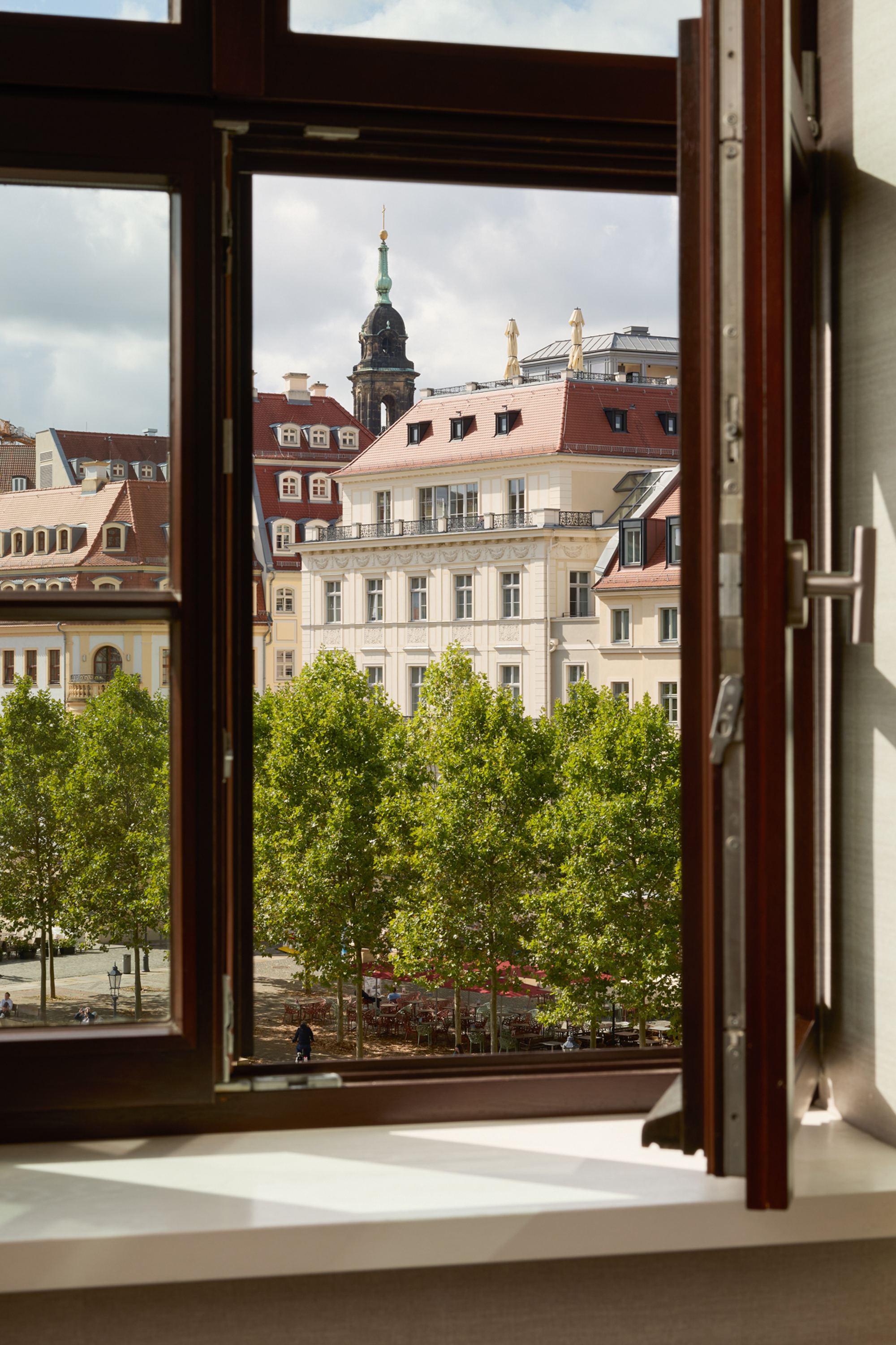 Blick aus offenem Hotelzimmerfenster auf historische Gebäude und grüne Bäume an einem städtischen Platz bei Tageslicht.