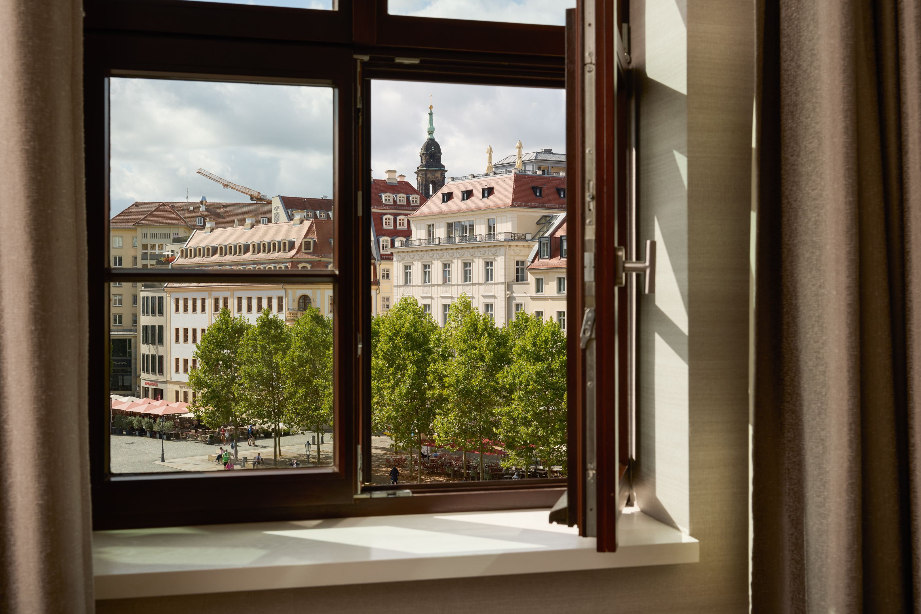 Blick aus offenem Hotelzimmerfenster auf historische Gebäude und grüne Bäume an einem städtischen Platz bei Tageslicht. Blick aus offenem Hotelzimmerfenster auf historische Gebäude und grüne Bäume an einem städtischen Platz bei Tageslicht.
