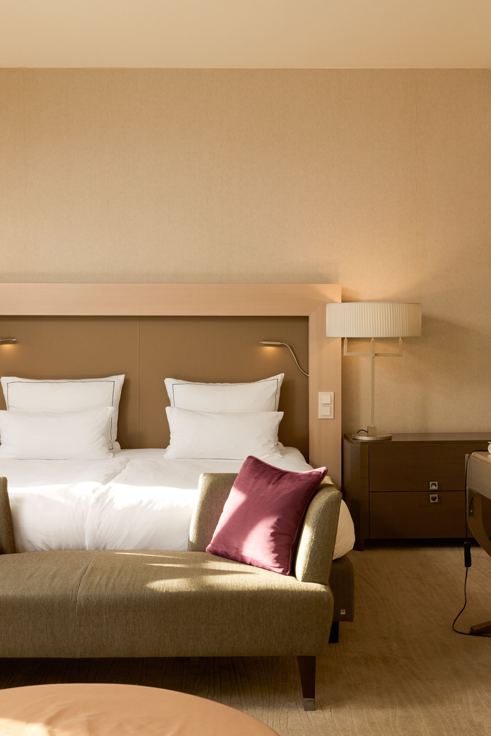Helles, modern eingerichtetes Hotelzimmer mit großem Doppelbett, zwei Kissen, einer gemütlichen Couch mit roten Zierkissen, Schreibtisch und warmem Tageslicht, das eine einladende und entspannte Atmosphäre schafft.