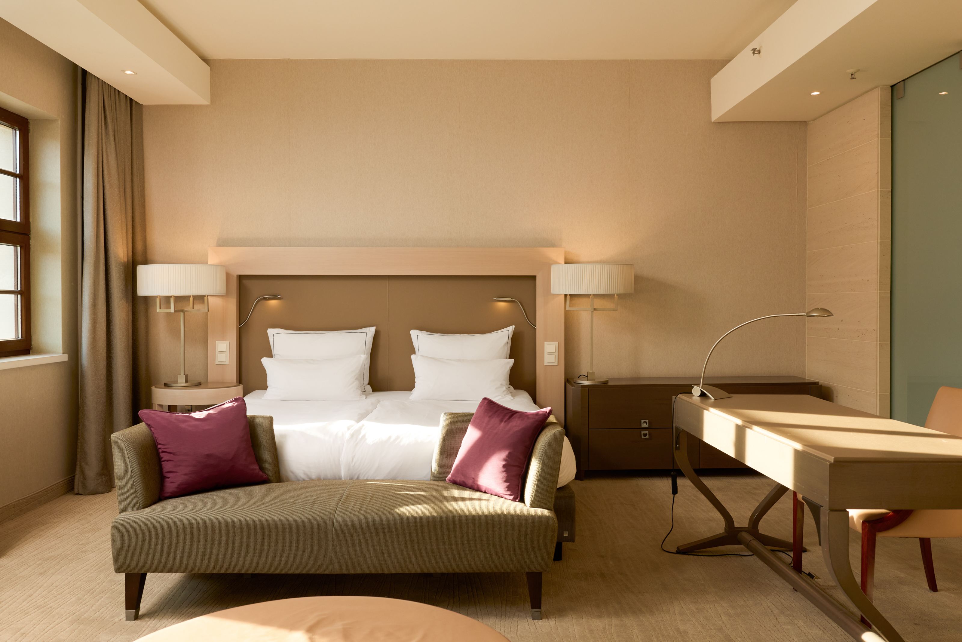 Helles, modern eingerichtetes Hotelzimmer mit großem Doppelbett, zwei Kissen, einer gemütlichen Couch mit roten Zierkissen, Schreibtisch und warmem Tageslicht, das eine einladende und entspannte Atmosphäre schafft. Helles, modern eingerichtetes Hotelzimmer mit großem Doppelbett, zwei Kissen, einer gemütlichen Couch mit roten Zierkissen, Schreibtisch und warmem Tageslicht, das eine einladende und entspannte Atmosphäre schafft.