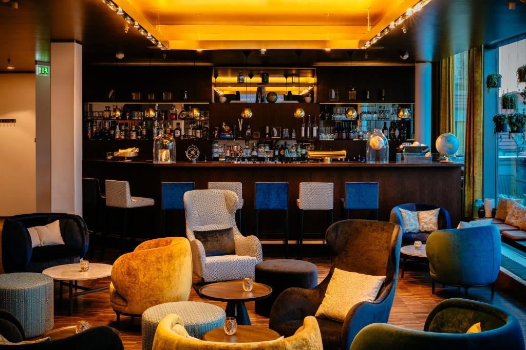 Gemütliche Hotelbar mit warmem Licht, bequemen Sesseln und kleinen Tischen, ideal zum Entspannen und geselligen Beisammensein. Elegante Bar im Hintergrund mit breitem Getränkeangebot. Gemütliche Hotelbar mit warmem Licht, bequemen Sesseln und kleinen Tischen, ideal zum Entspannen und geselligen Beisammensein. Elegante Bar im Hintergrund mit breitem Getränkeangebot.