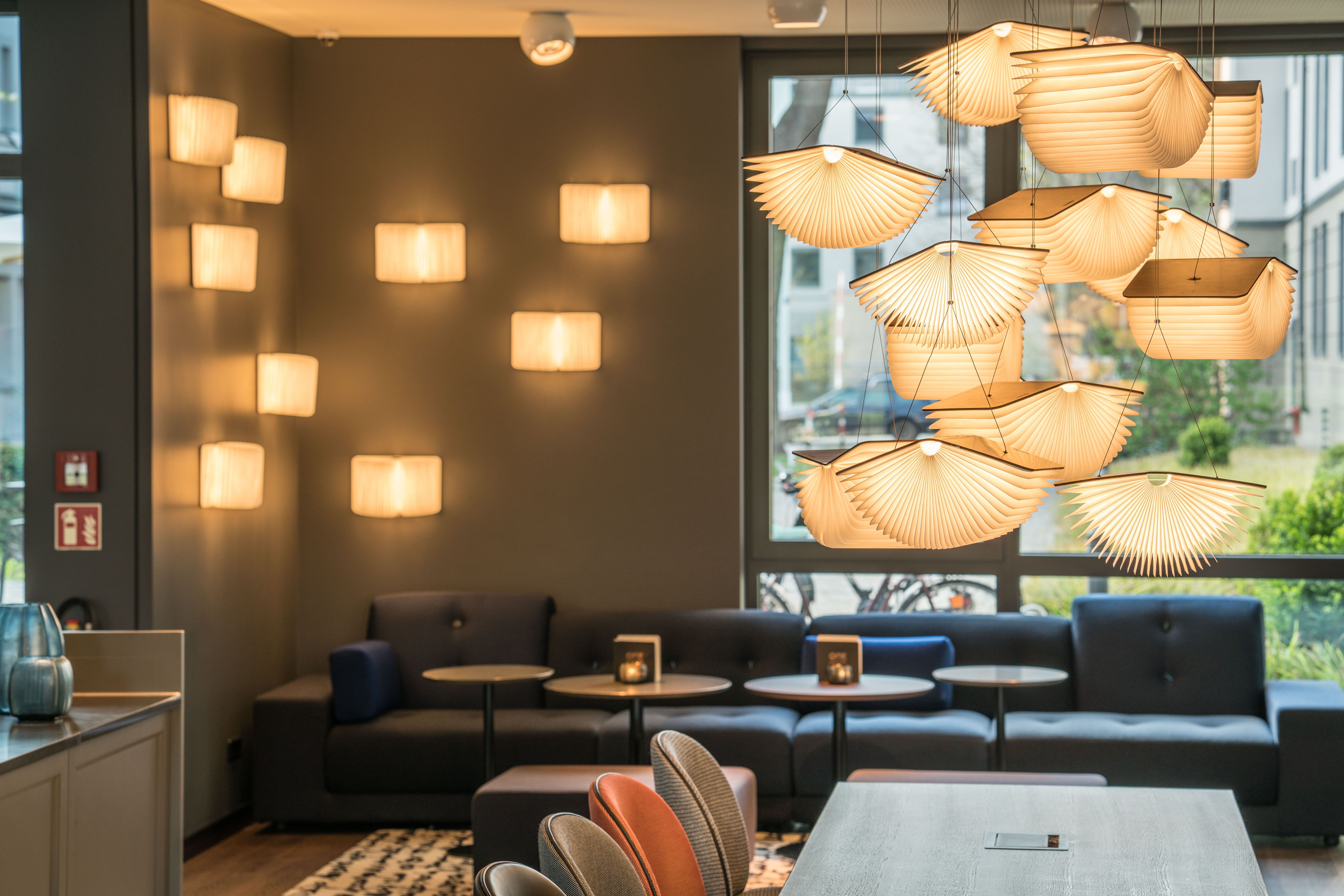 hotel Dresden Palaisplatz Motel One bright lounge with a long sofa in front of floor-to-ceiling windows, several ceiling lamps, and a table in the foreground