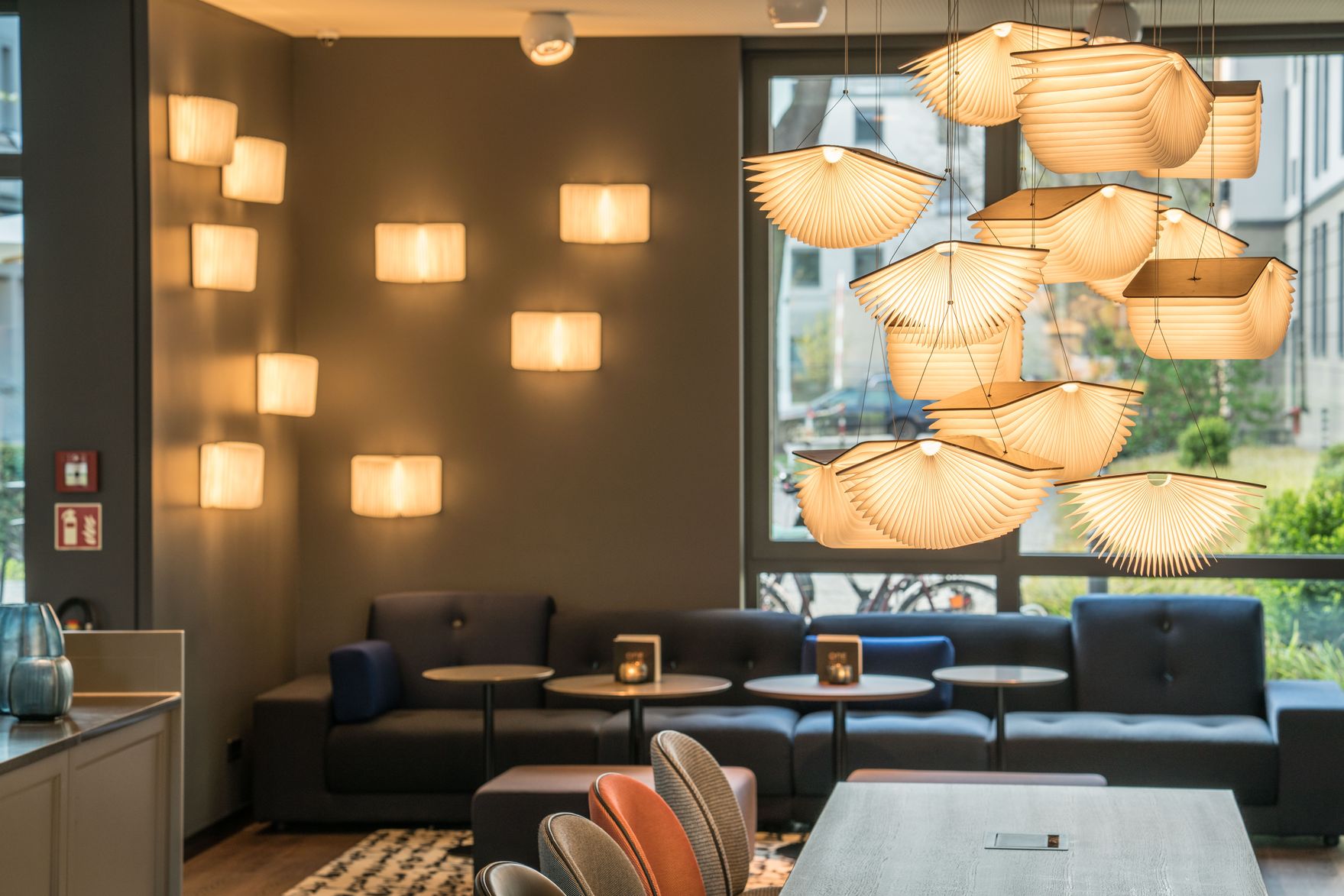 Hotel Dresden Palaisplatz Motel One helle Lounge mit langem Sofa vor bodentiefem Fenster, mehrere Deckenlampen sowie ein Tisch im Vordergrund