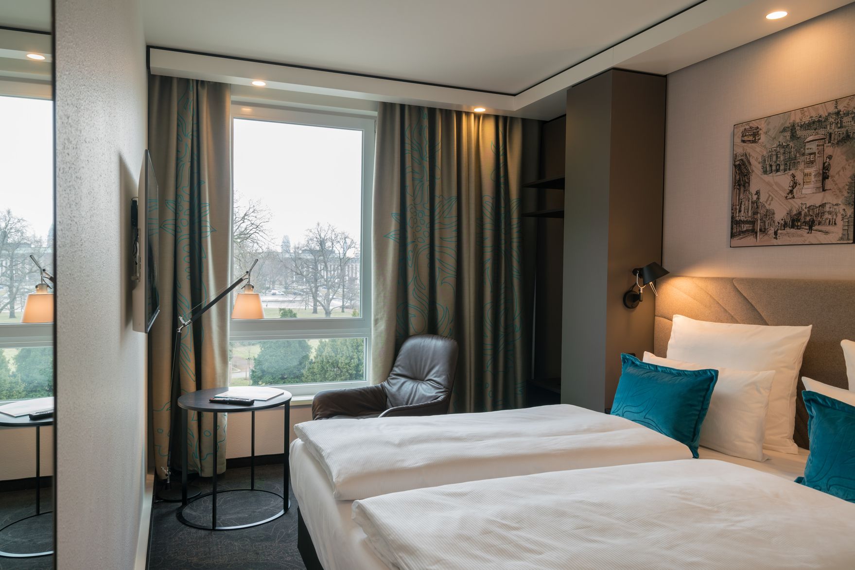 Designhotel Dresden Palaisplatz Motel One seitlicher Blick in lichtdurchflutetes Zimmer mit Doppelbet, Sessel, Beistelltisch sowie TV an der Wand