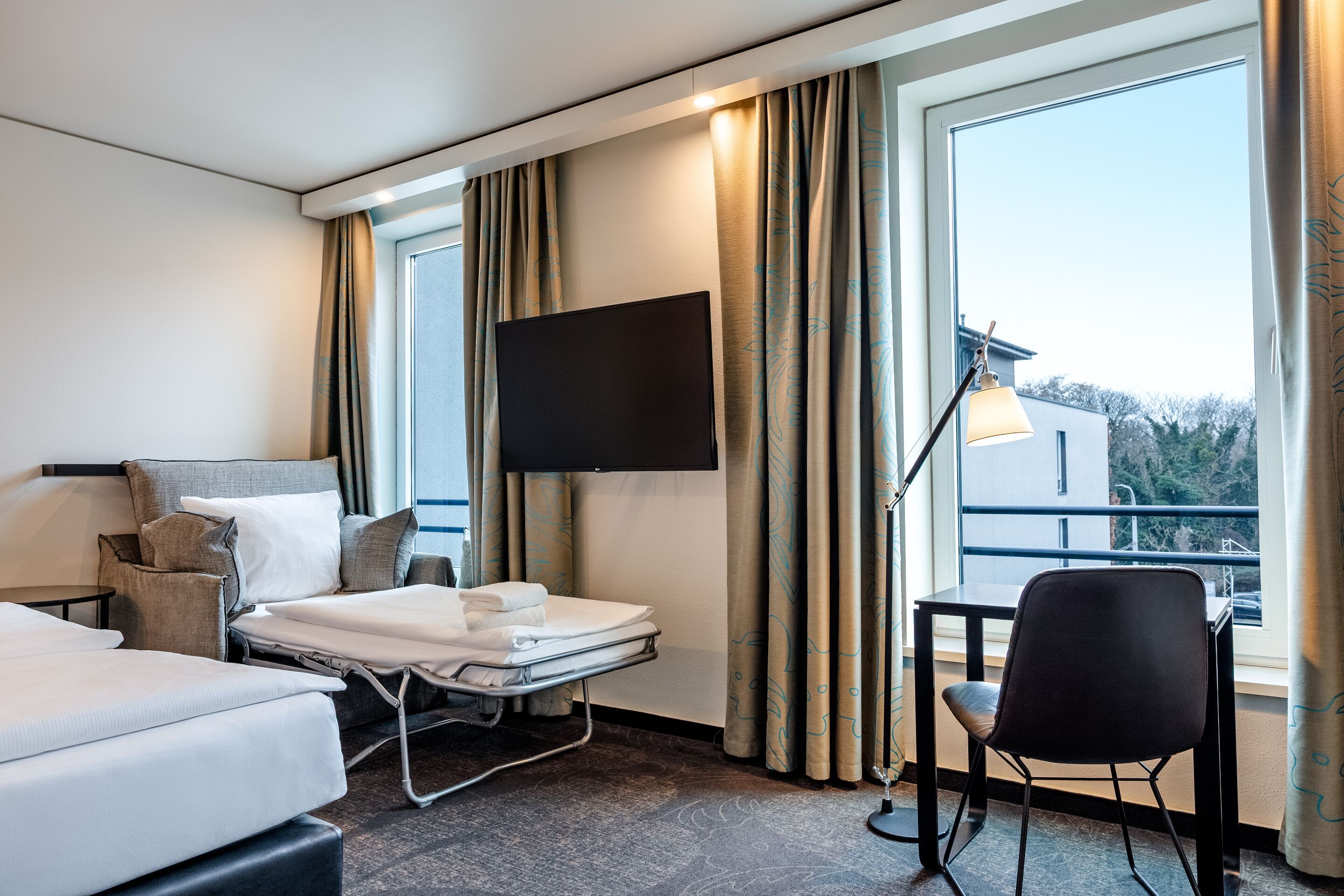 Hotel Motel One Dresden-Palaisplatz Helles Hotelzimmer mit zwei Betten, einem ausziehbaren Gästebett, Fenster mit Blick ins Grüne, Schreibtisch mit Stuhl und Leselampe sowie einem an der Wand montierten Fernseher. Vorhänge rahmen die Fenster und schaffen eine gemütliche Atmosphäre.