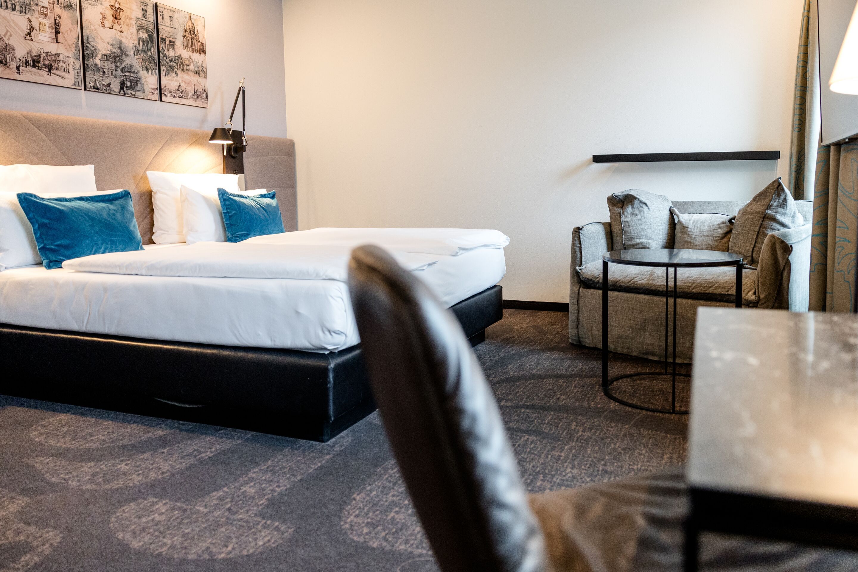 Hotel Motel One Dresden-Palaisplatz Modern eingerichtetes Hotelzimmer mit großem Doppelbett, hellen Wänden, drei blauen Kissen, Sitzbereich mit zwei Sesseln und kleinem runden Tisch, angenehmer, ruhiger Atmosphäre.