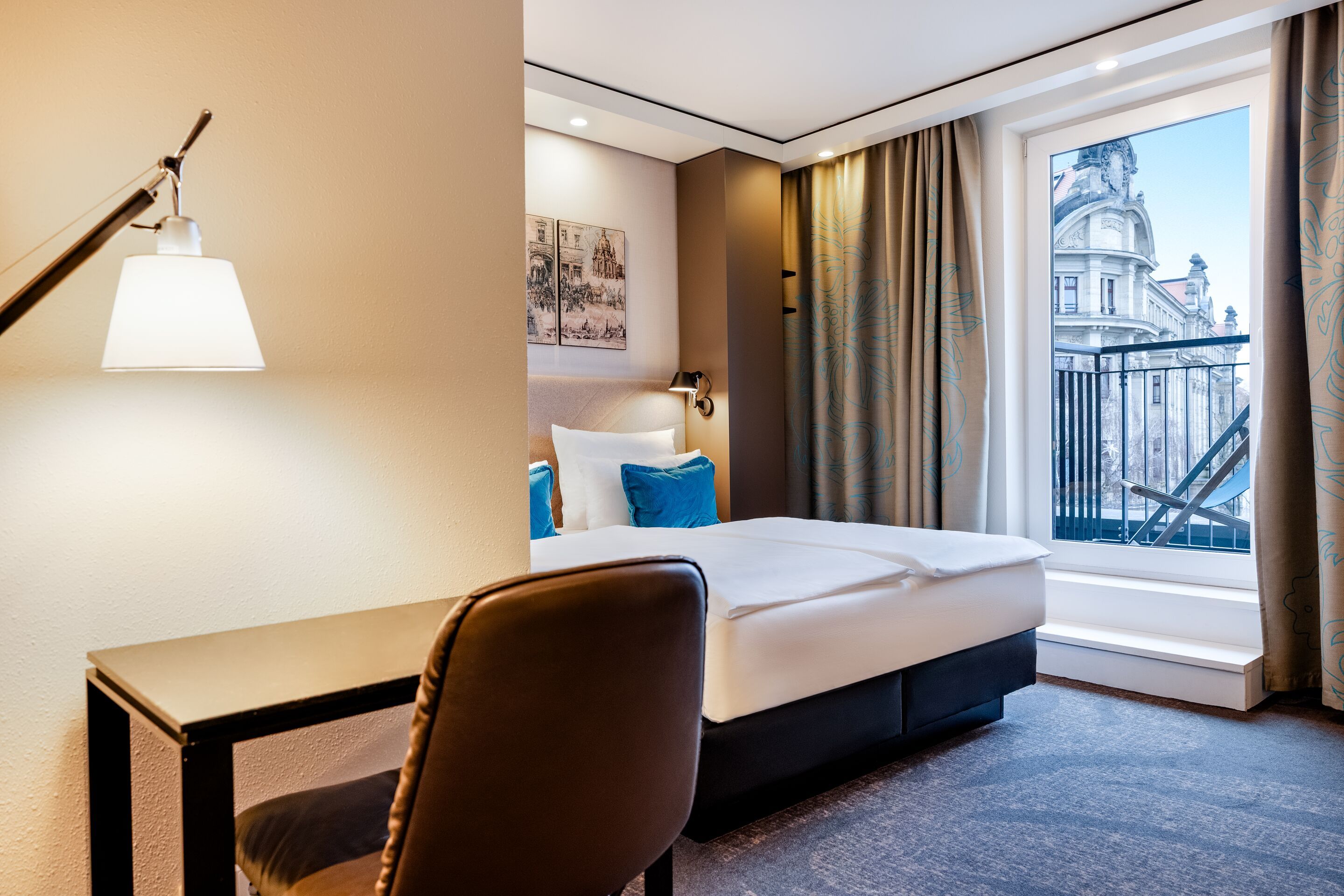 Hotel Motel One Dresden-Palaisplatz Modern eingerichtetes Hotelzimmer mit Einzelbett, Schreibtisch und Sitzgelegenheit. Große Fensterfront mit Blick auf historische Gebäude. Helle, gemütliche Atmosphäre durch warmes Licht und dezente Dekoration. Ideal für Geschäftsreisende und Städteurlauber.