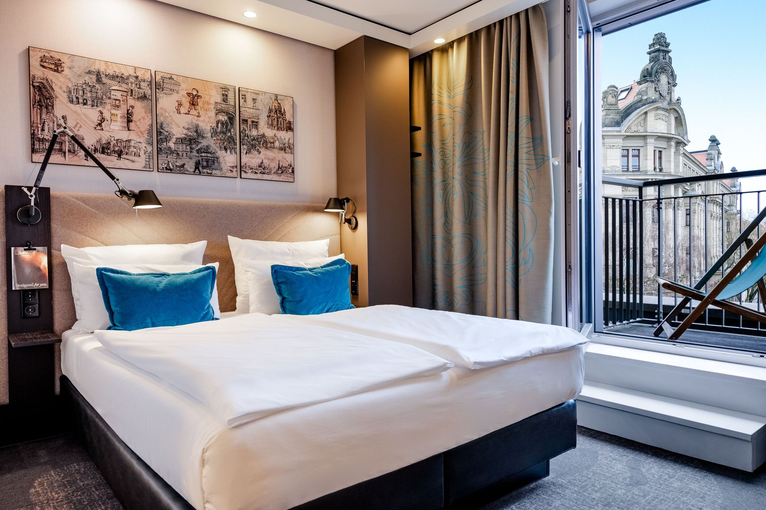 Modernes Hotelzimmer mit großem, bequemem Doppelbett, blauen Akzentkissen und stimmungsvoller Beleuchtung. Balkon mit Sitzgelegenheit und Blick auf historische Architektur im Stadtzentrum. Helle, freundliche Atmosphäre für erholsamen Aufenthalt.