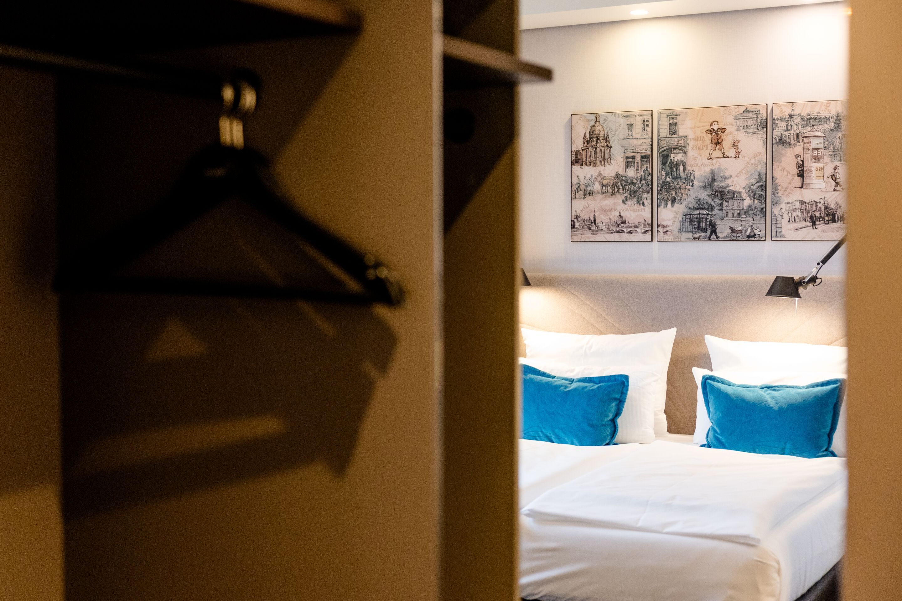 Hotel Motel One Dresden-Palaisplatz Ein Blick in ein modernes, gemütliches Hotelzimmer mit einem großen, weißen Bett, blauen Kissen und Wandbildern. Im Vordergrund ein offener Kleiderschrank mit schwarzen Kleiderbügeln. Warme Beleuchtung sorgt für eine einladende Atmosphäre.