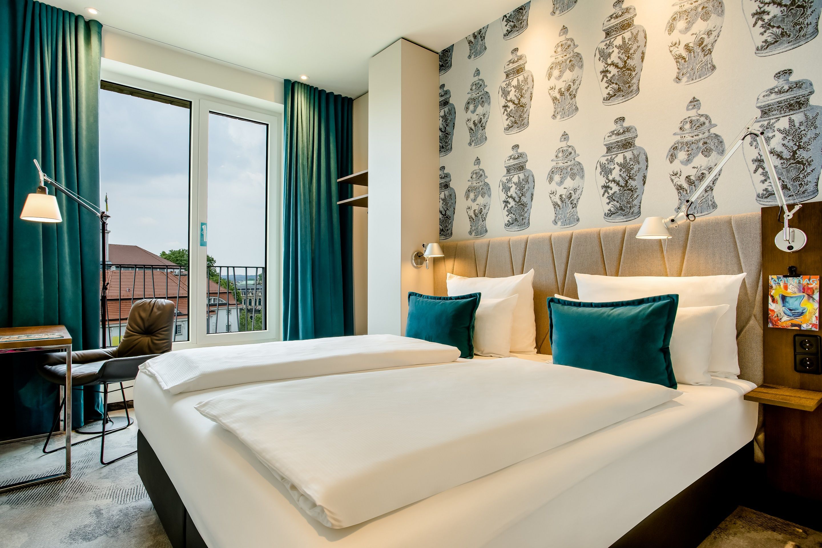 Designhotel Dresden am Zwinger Motel One Blick auf großes Doppelbett mit zwei Zierkissen in türkis sowie Tisch und Stuhl vor bodentiefem Fenster