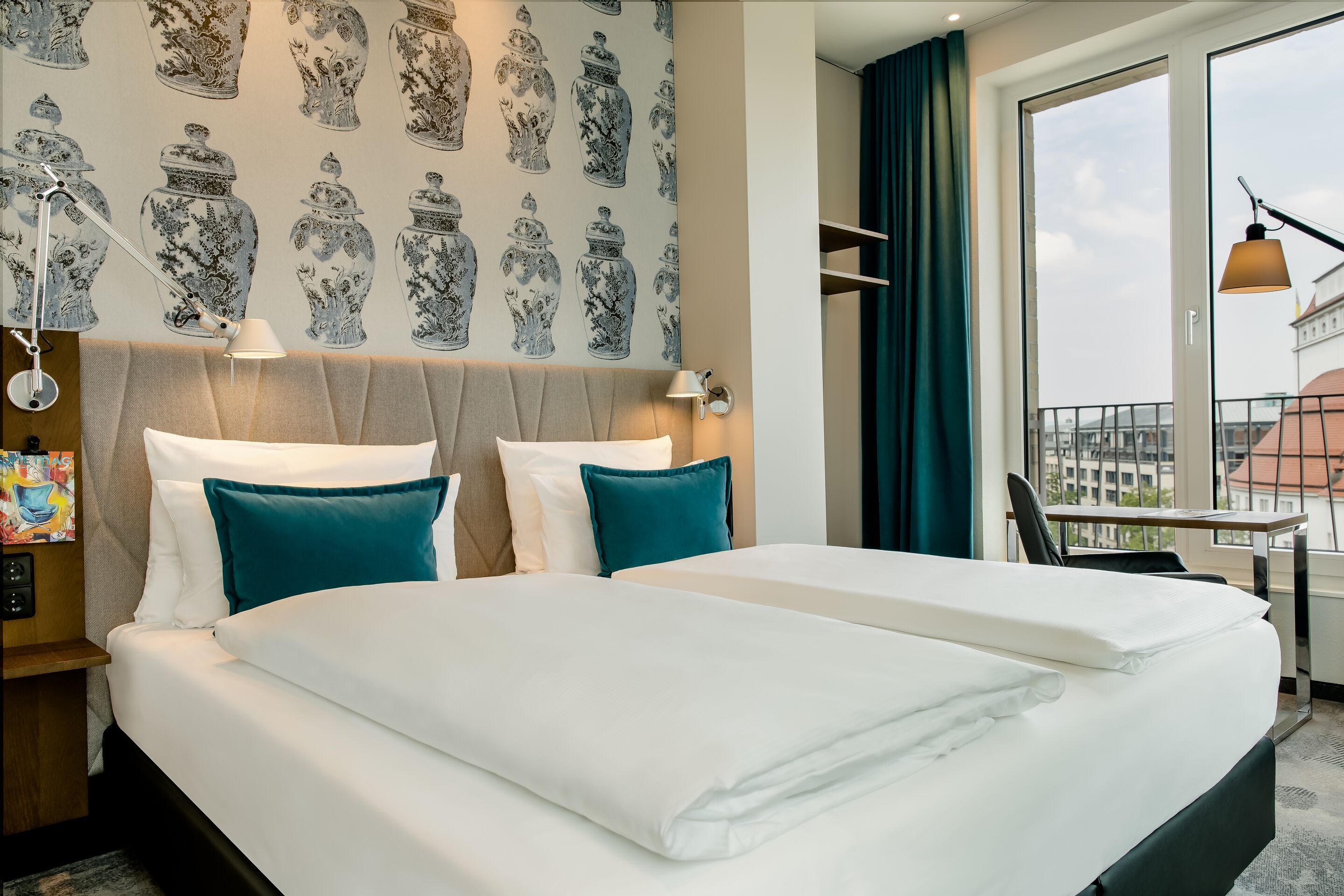 Hotel Motel One Dresden am Zwinger Modern eingerichtetes Hotelzimmer mit großem Doppelbett, weißen Bettdecken und türkisfarbenen Kissen. Große Fenster bieten Stadtblick, ein Schreibtisch und dezente Wanddekoration schaffen eine einladende Atmosphäre.