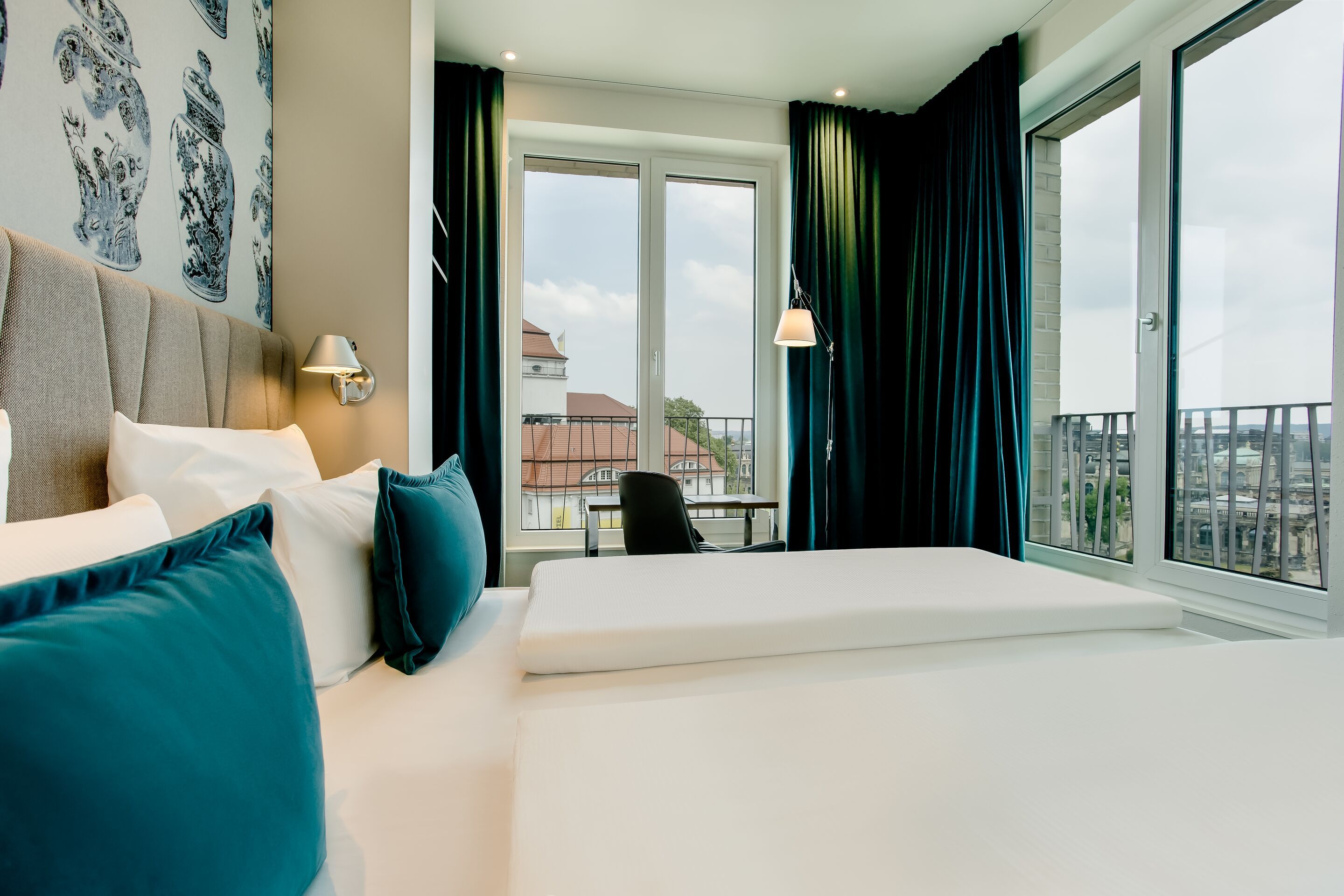 Hotel Motel One Dresden am Zwinger Helles Hotelzimmer mit großem gemütlichem Bett, blauen Kissen, bodentiefen Fenstern und Blick auf eine Stadtlandschaft. Moderner, ruhiger Raum mit Schreibtisch und Vorhängen für entspannten Aufenthalt.