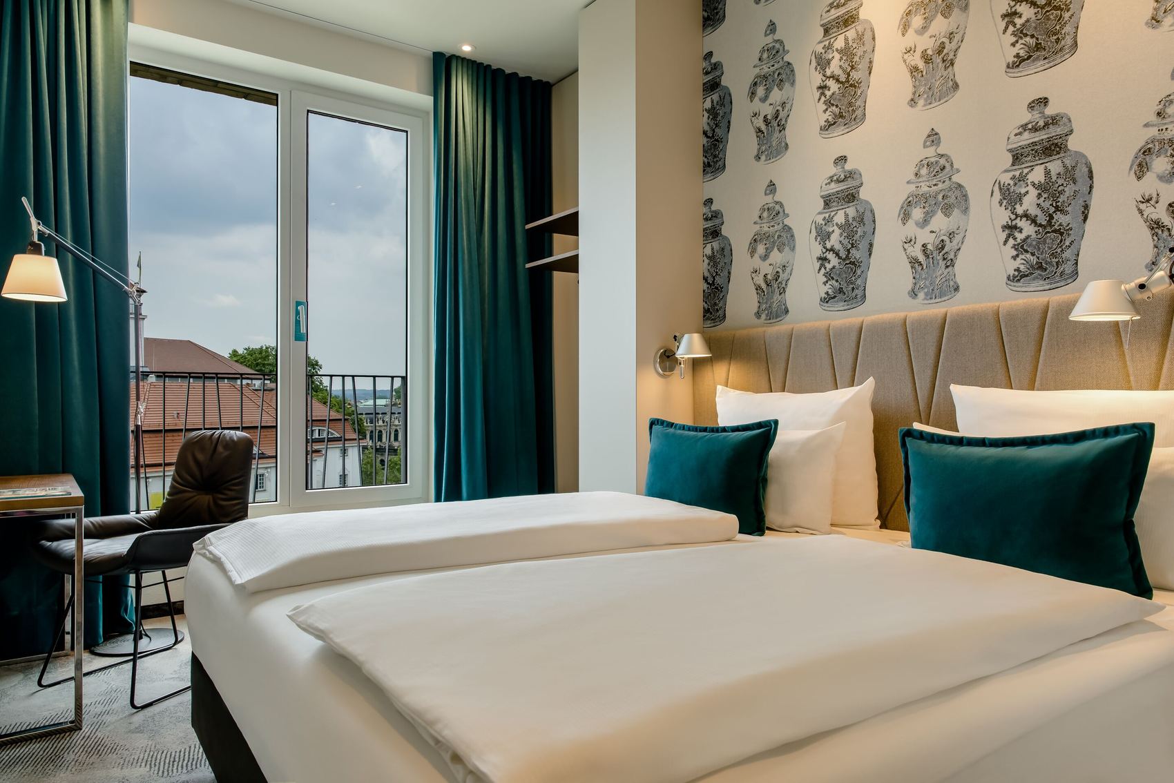 Hotel Motel One Dresden am Zwinger Modern eingerichtetes Hotelzimmer mit großem Bett, Akzentkissen in Blau, stilvoller Tapete und Balkon mit Ausblick auf Stadtgebäude, helle und ruhige Atmosphäre.