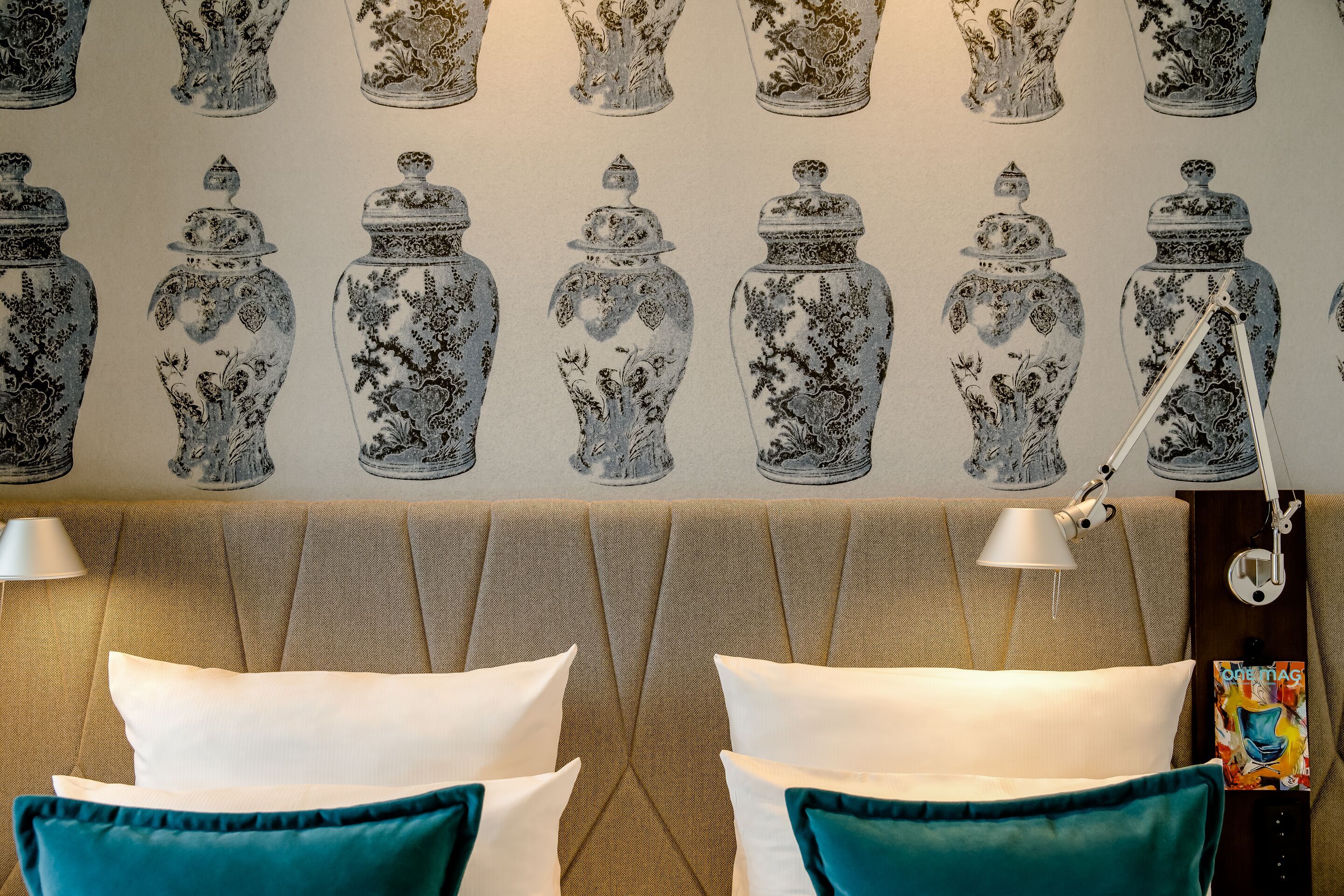 Hotel Motel One Dresden am Zwinger Beidseitiges Doppelbett mit weißen Kissen und türkisfarbenen Zierkissen vor gepolsterter Rückwand. Wand mit dekorativem Tapetenmuster aus schwarz-weißen Vasenmotiven. Zwei verstellbare Leselampen an der Wand. Stimmungsvoll und modern eingerichtet.