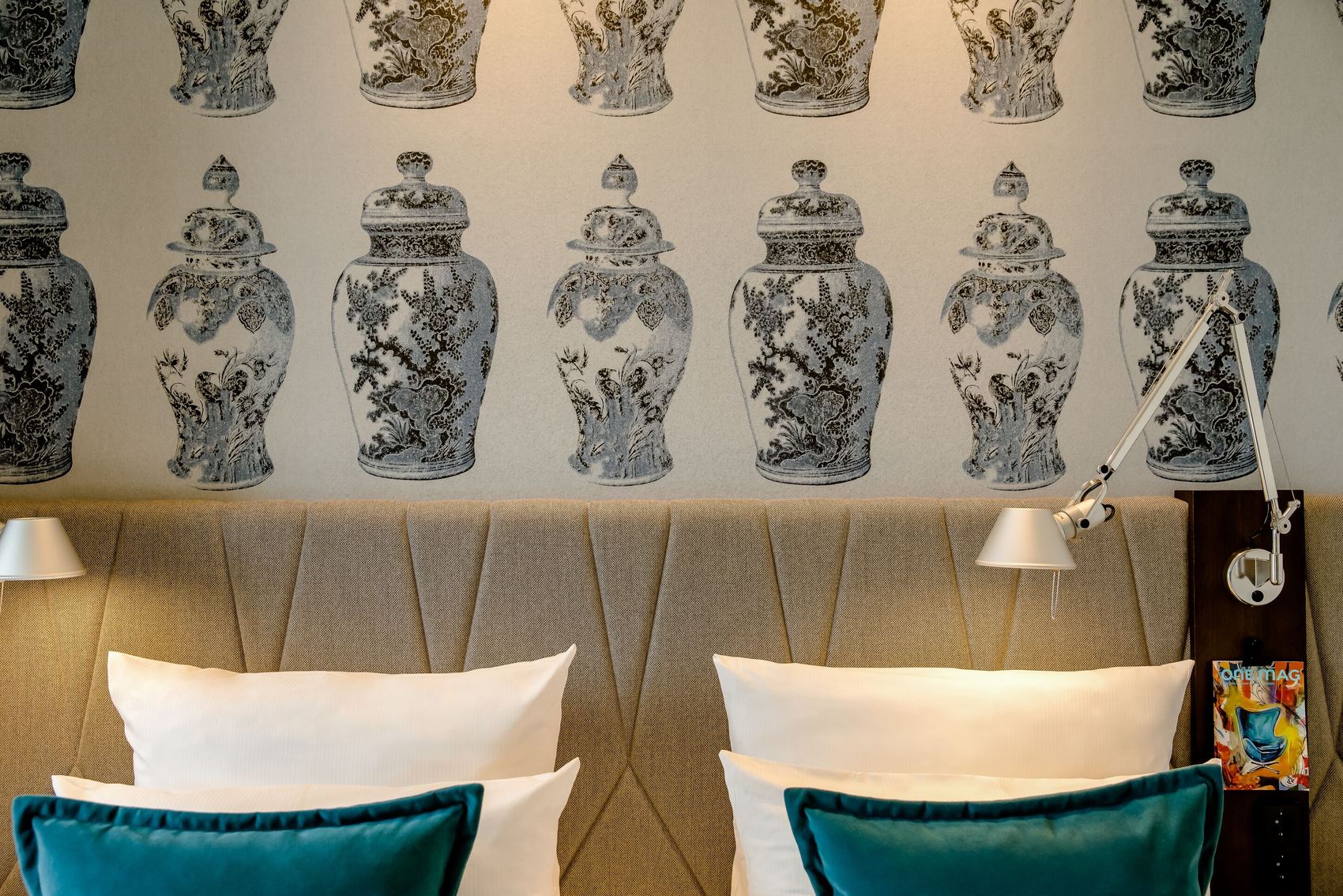 Hotel Motel One Dresden am Zwinger Beidseitiges Doppelbett mit weißen Kissen und türkisfarbenen Zierkissen vor gepolsterter Rückwand. Wand mit dekorativem Tapetenmuster aus schwarz-weißen Vasenmotiven. Zwei verstellbare Leselampen an der Wand. Stimmungsvoll und modern eingerichtet.