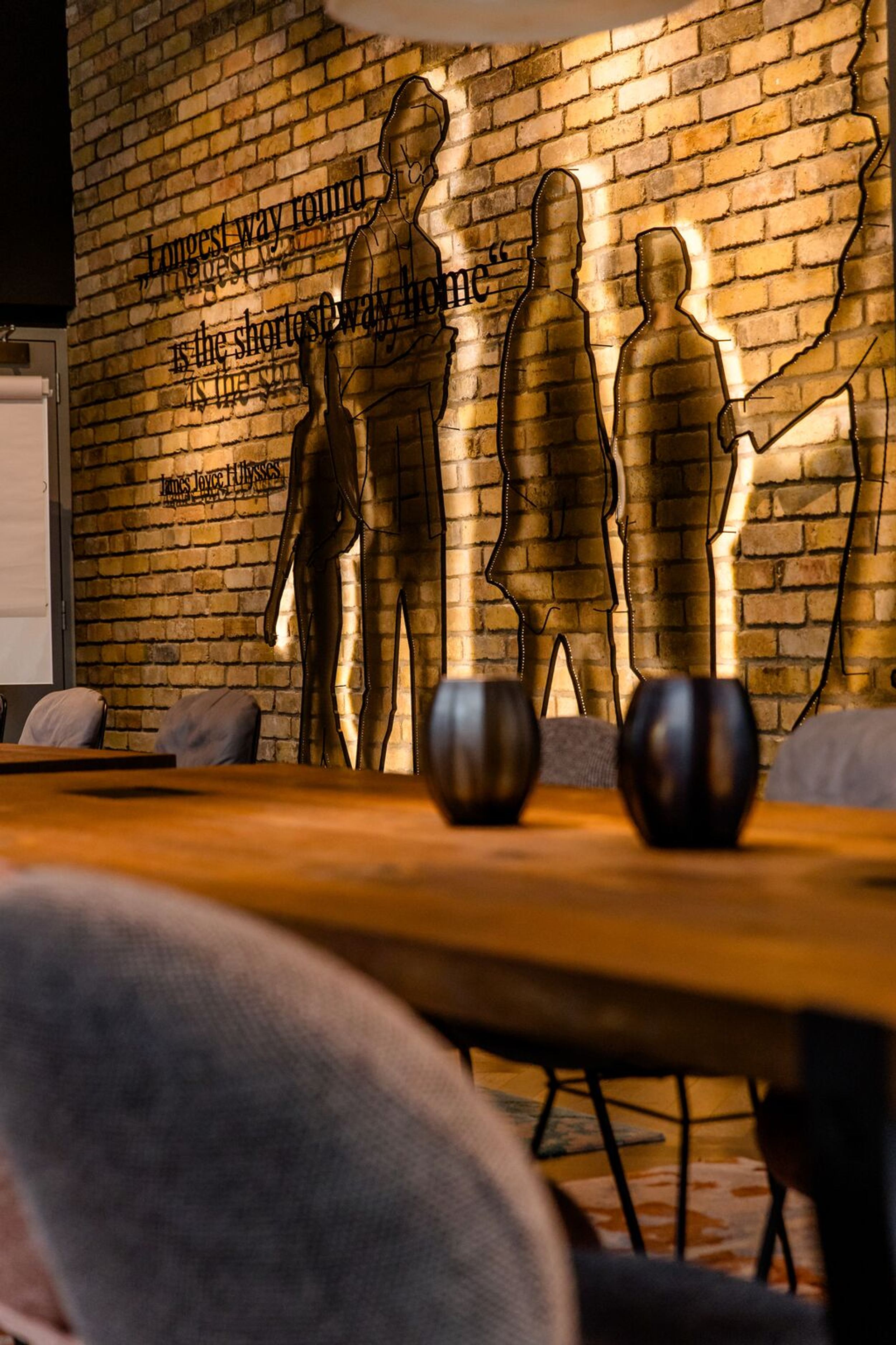 Hotel Dublin Motel One langer Tisch mit Stühlen vor Wandinstallation mit Umrissen von Personen sowie einem Zitat von James Joyce