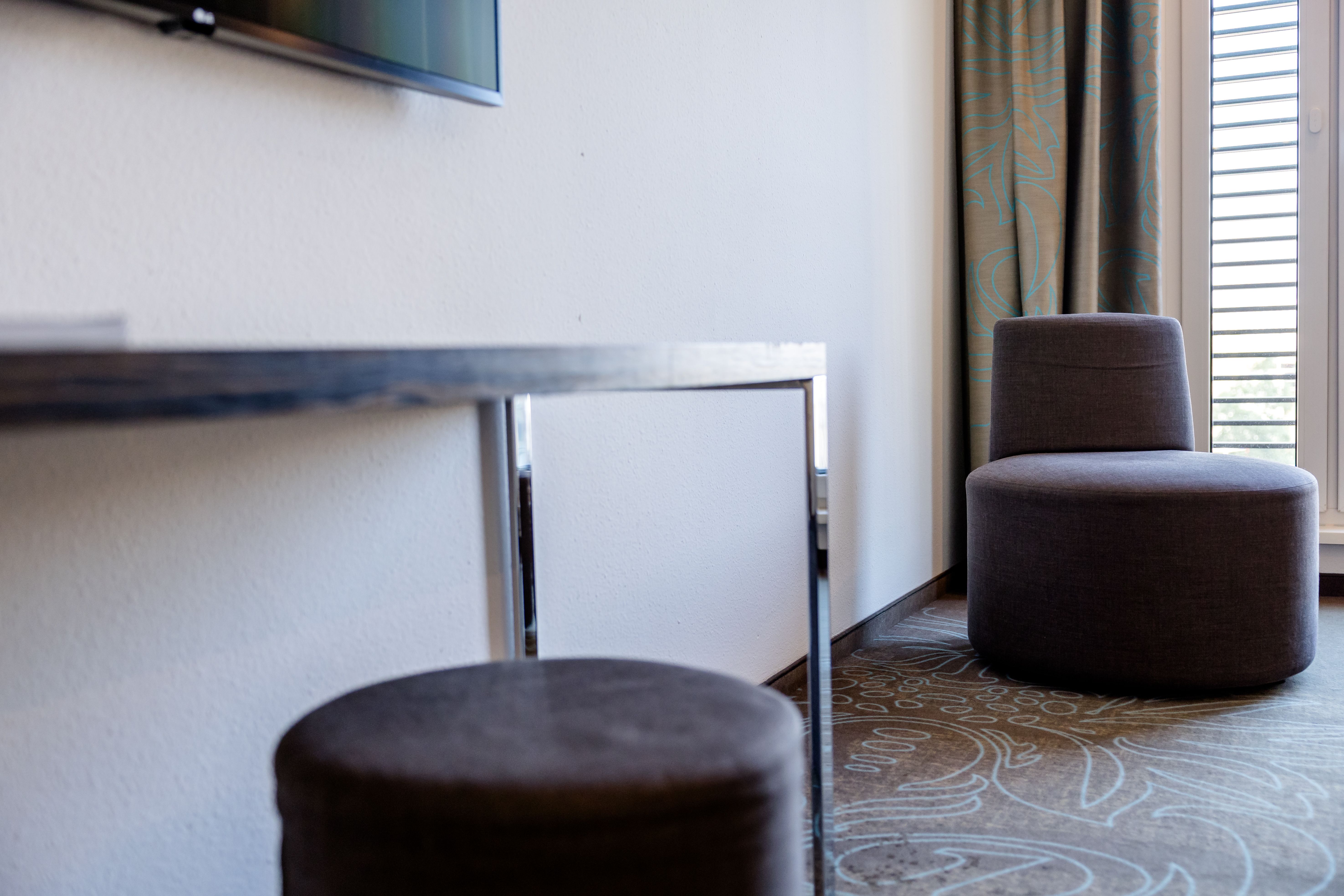 Moderner Hotelzimmerbereich mit gemütlichem, runden Sessel und Hocker in neutralen Brauntönen, hellem Teppich mit floralem Muster, lichtdurchflutet durch Fenster mit halb geöffnetem Vorhang.