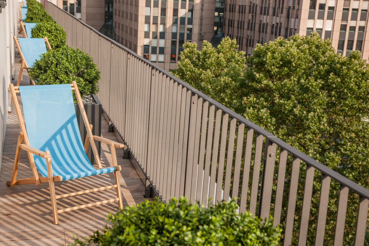 Balkon mit mehreren blauen Liegestühlen und grünen Pflanzen, Lärmschutz durch Balkongeländer, im Hintergrund Hochhäuser und Bäume.