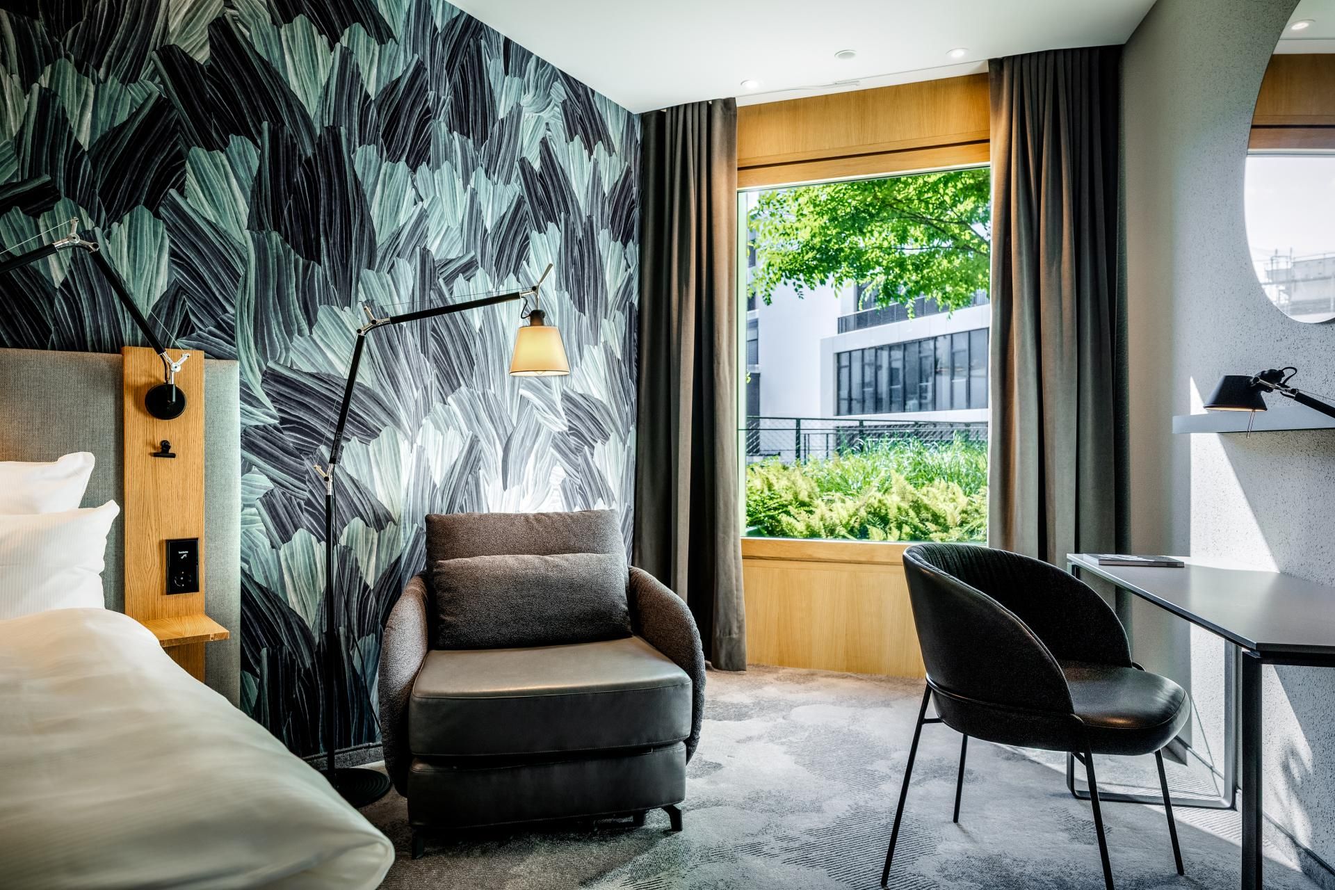 Modern eingerichtetes Hotelzimmer mit bequemen Sessel, Schreibtisch und großem Fenster, das viel Tageslicht und Blick auf grüne Pflanzen bietet. Die Wandgestaltung mit Blattmotiv verleiht dem Raum eine natürliche, entspannte Atmosphäre.