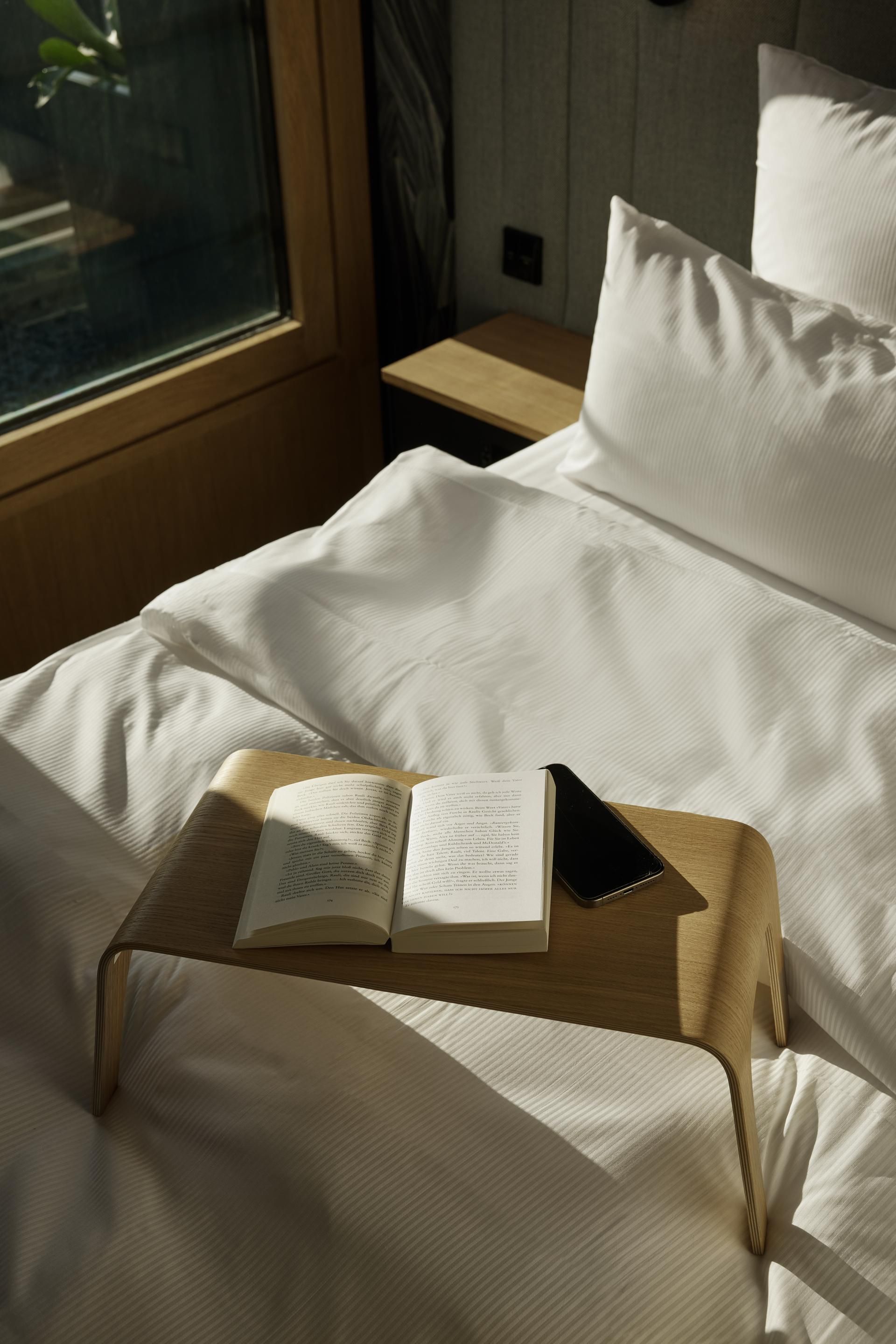 Ein helles, gemütliches Bett mit weißen Bettwäsche, darauf ein kleiner Holztisch mit einem geöffneten Buch und einem Smartphone. Sonnenlicht fällt durch das Fenster und schafft eine entspannte Atmosphäre.