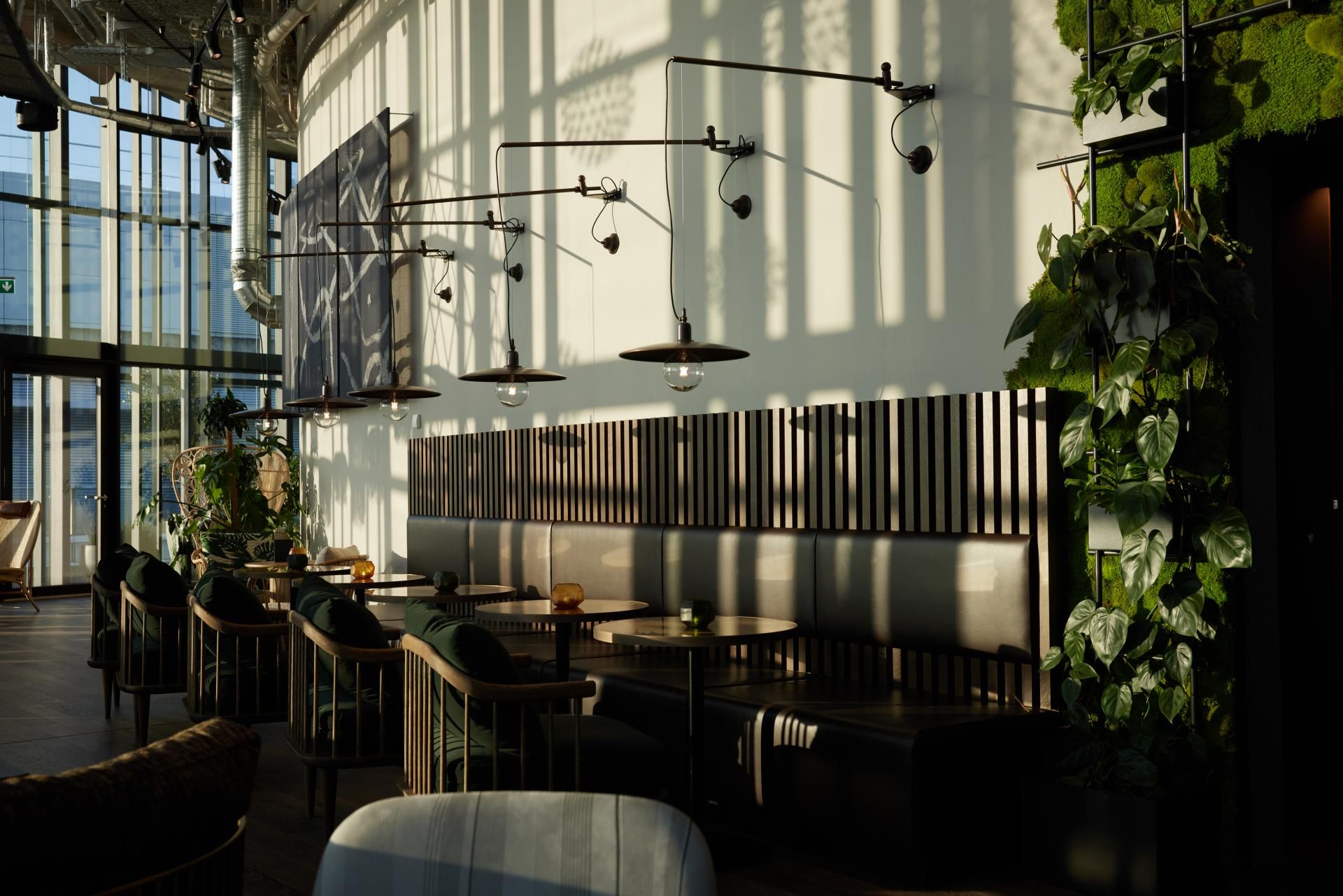 Heller, moderner Restaurantbereich mit hohen Fenstern, stilvollen Tischen und Stühlen, warmem Sonnenlicht und grünen Pflanzen an den Wänden für eine einladende Atmosphäre.