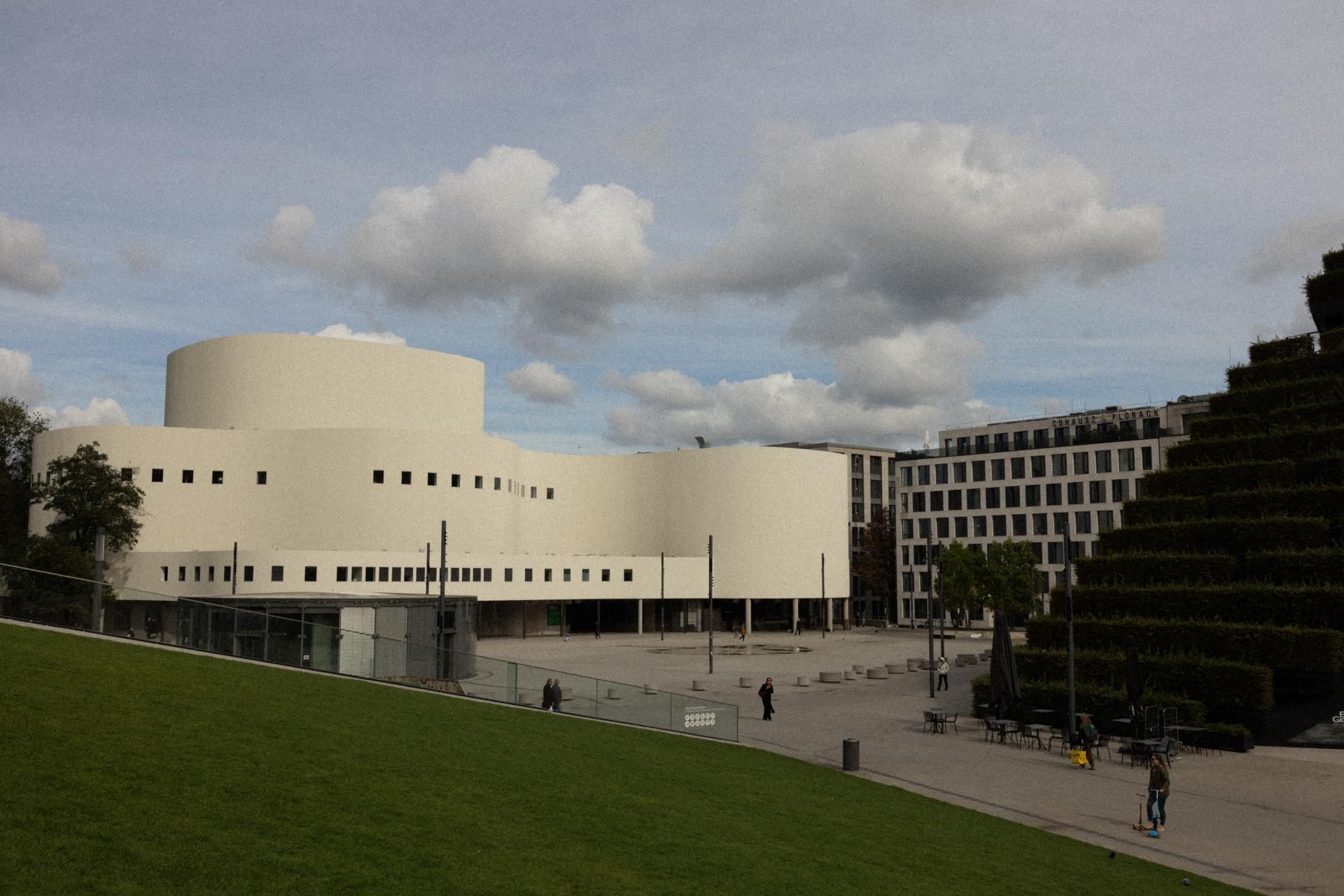 Modernes, weißes Veranstaltungsgebäude mit runden Formen und vielen kleinen Fenstern, umgeben von offenem Platz und grünen Flächen unter bewölktem Himmel, vermittelt ein urbanes, ruhiges Ambiente.