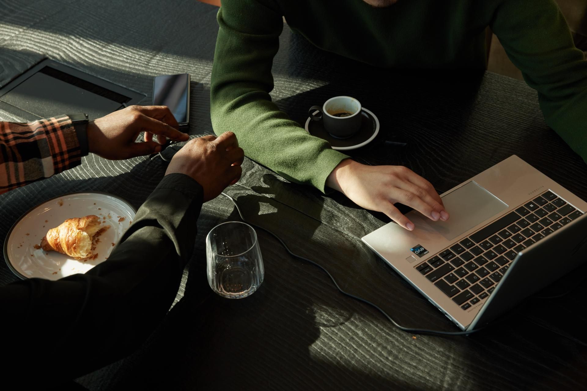 Zwei Personen an einem Tisch mit Laptop, Kaffee und Croissant, eine Person tippt auf der Tastatur, Atmosphäre wirkt entspannt und konzentriert.